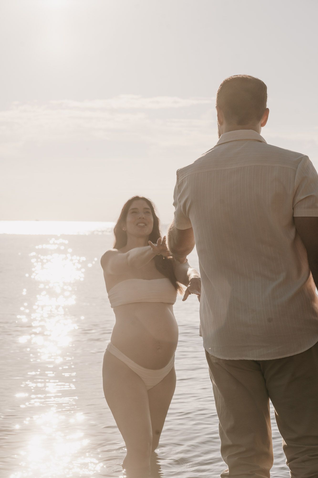 Pregnant woman in white bikini holding hands with man while wading in sparkling sunlit ocean water.