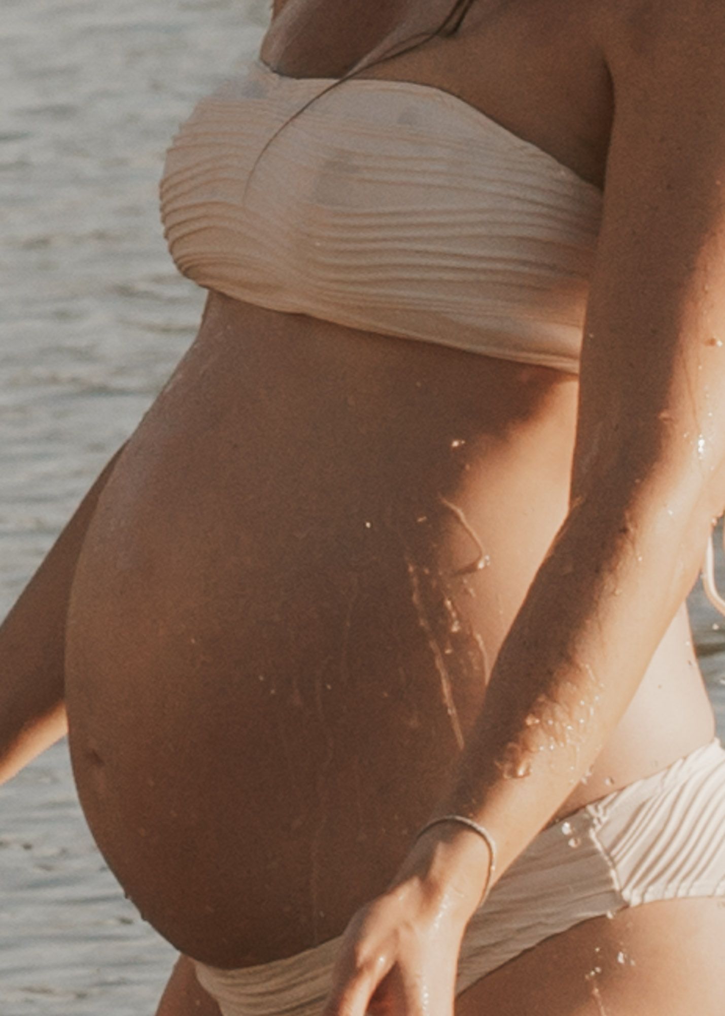 Pregnant woman in a beige bandeau bikini showing baby bump at the beach in warm sunlight.