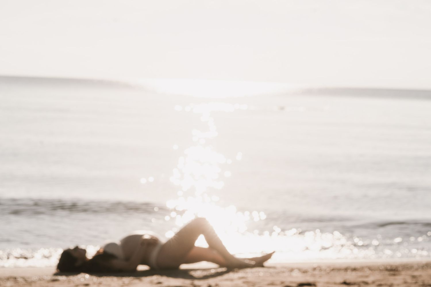 Woman in bikini lying on sandy beach with bright sunlight reflecting off the ocean in the background.