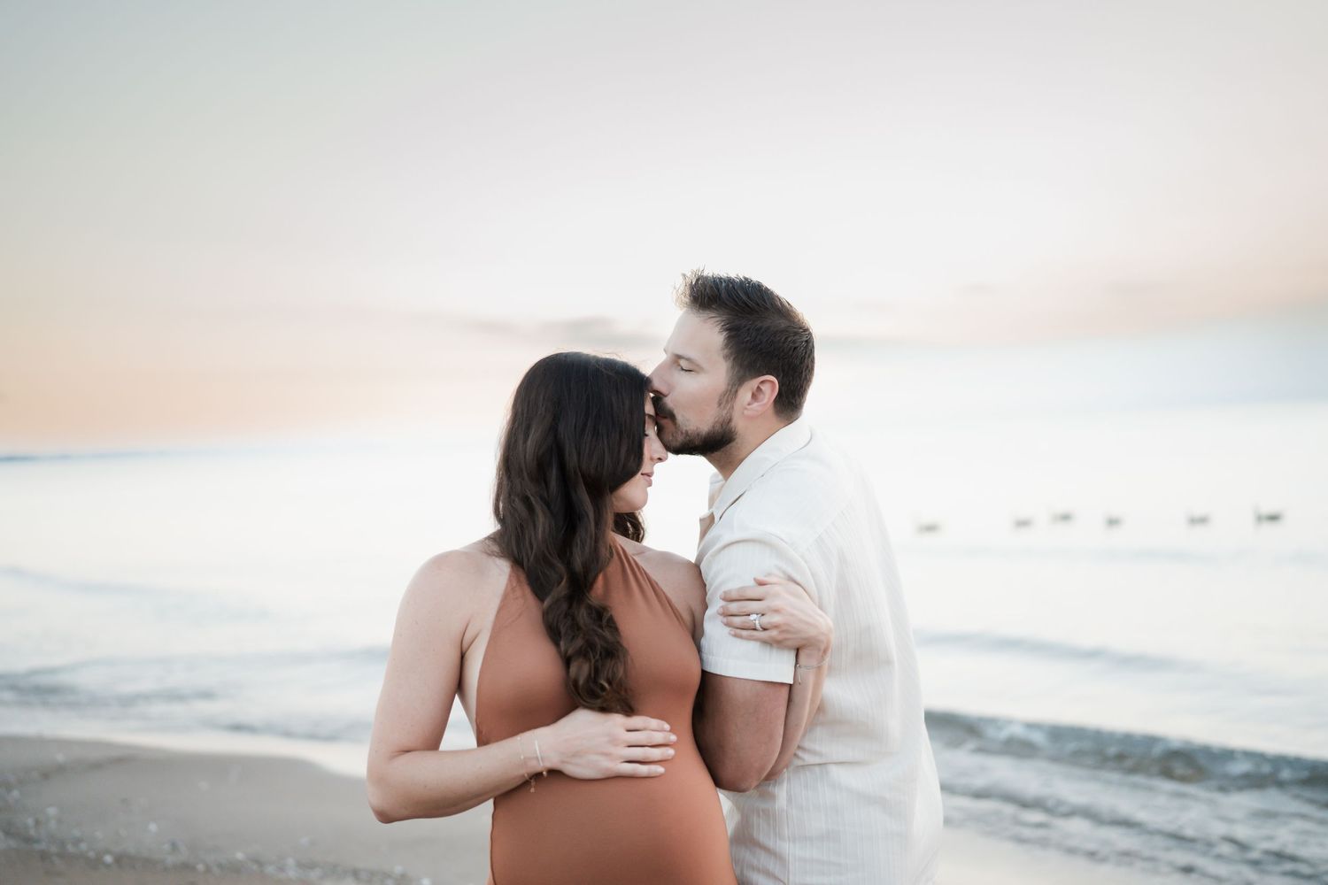 Couple sharing a tender kiss on the beach at sunset, with the woman in a rust-colored maternity dress.