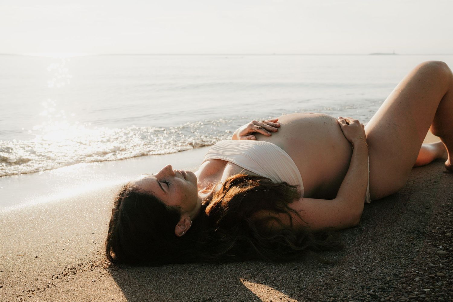 Pregnant woman in white bikini lying on sandy beach with ocean waves and soft sunlight in the background.