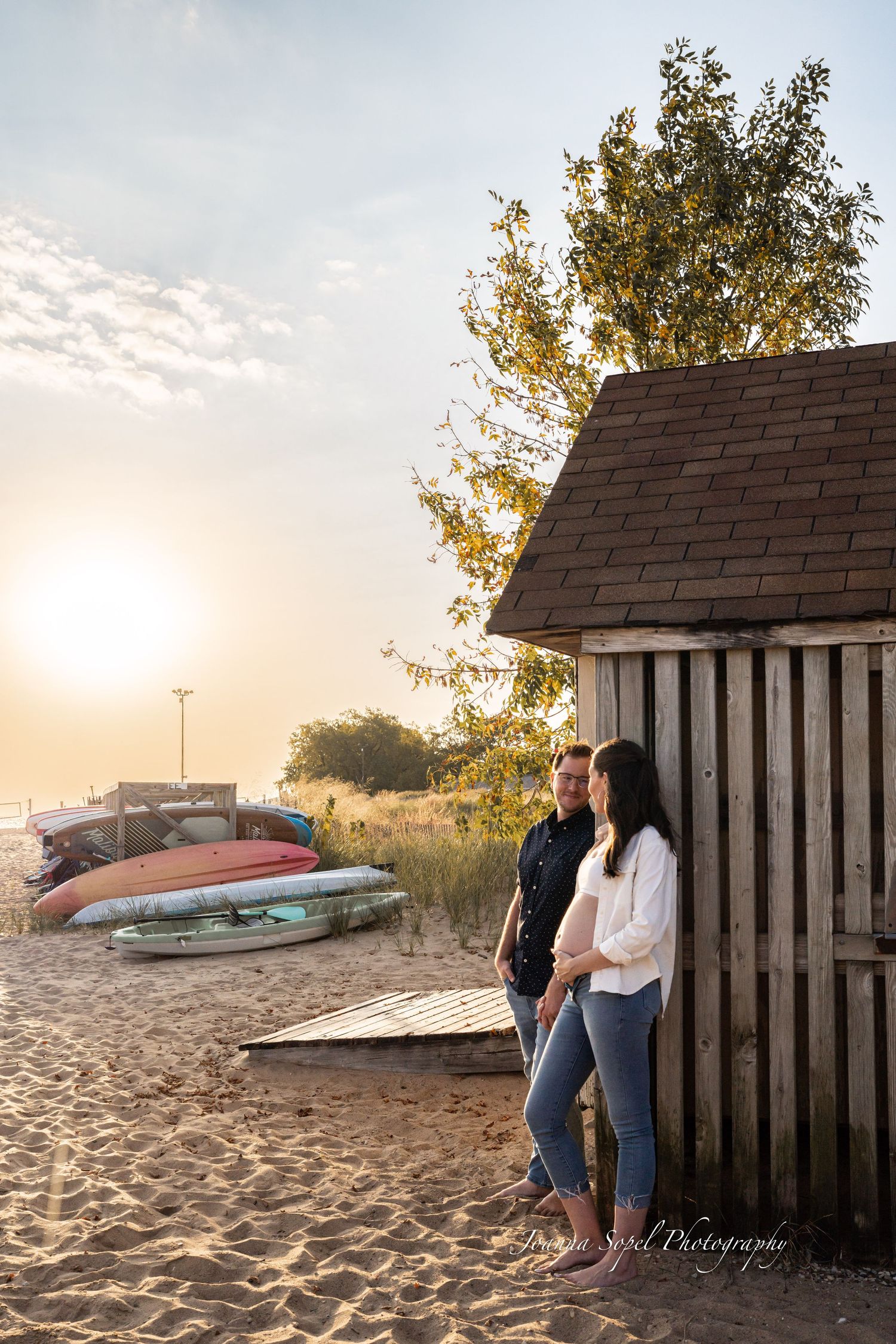 Couple standing by rustic beach hut at golden sunset with colorful kayaks nearby.