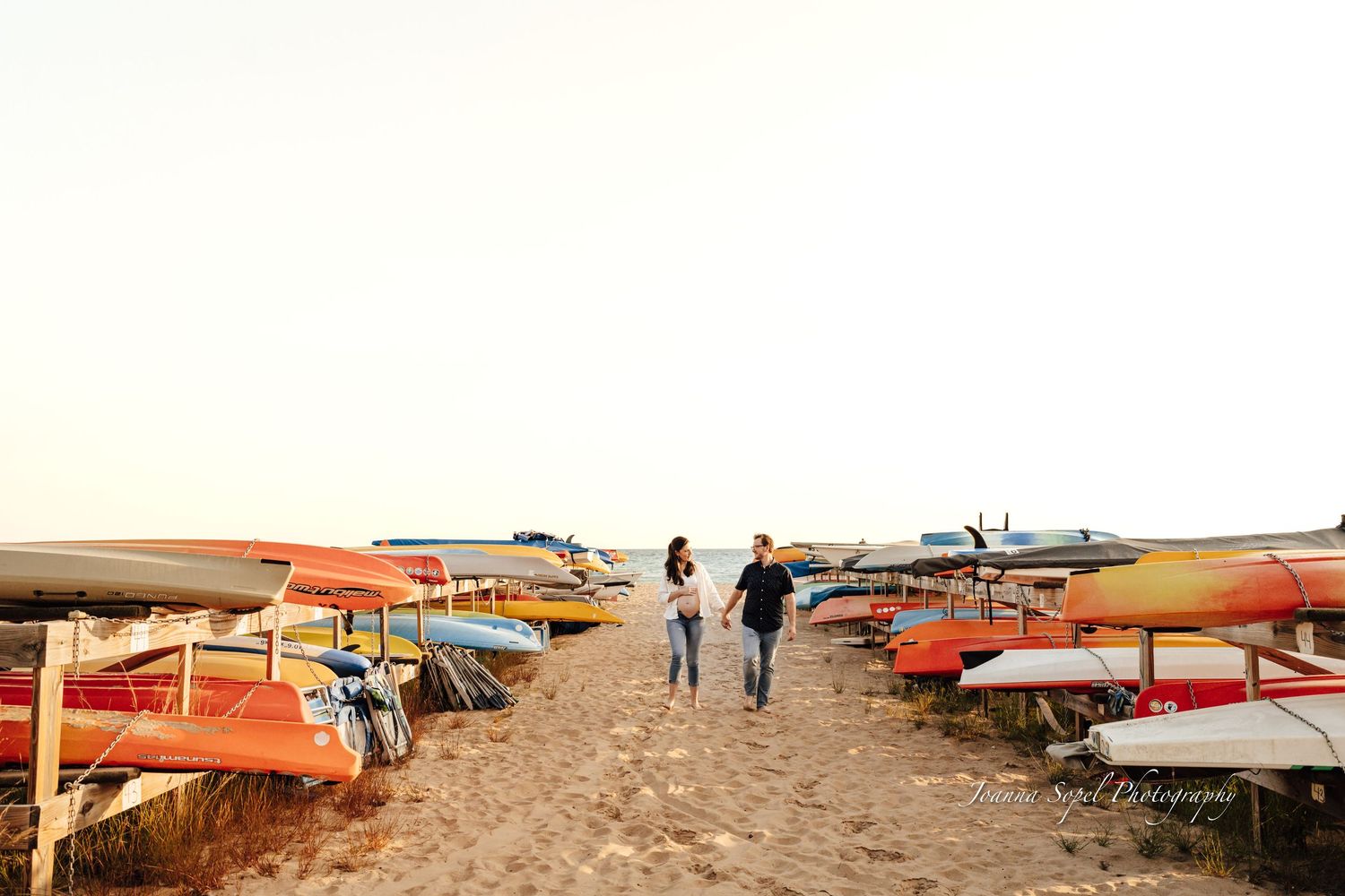 Romantic couple walking hand in hand between rows of colorful kayaks on sandy beach.