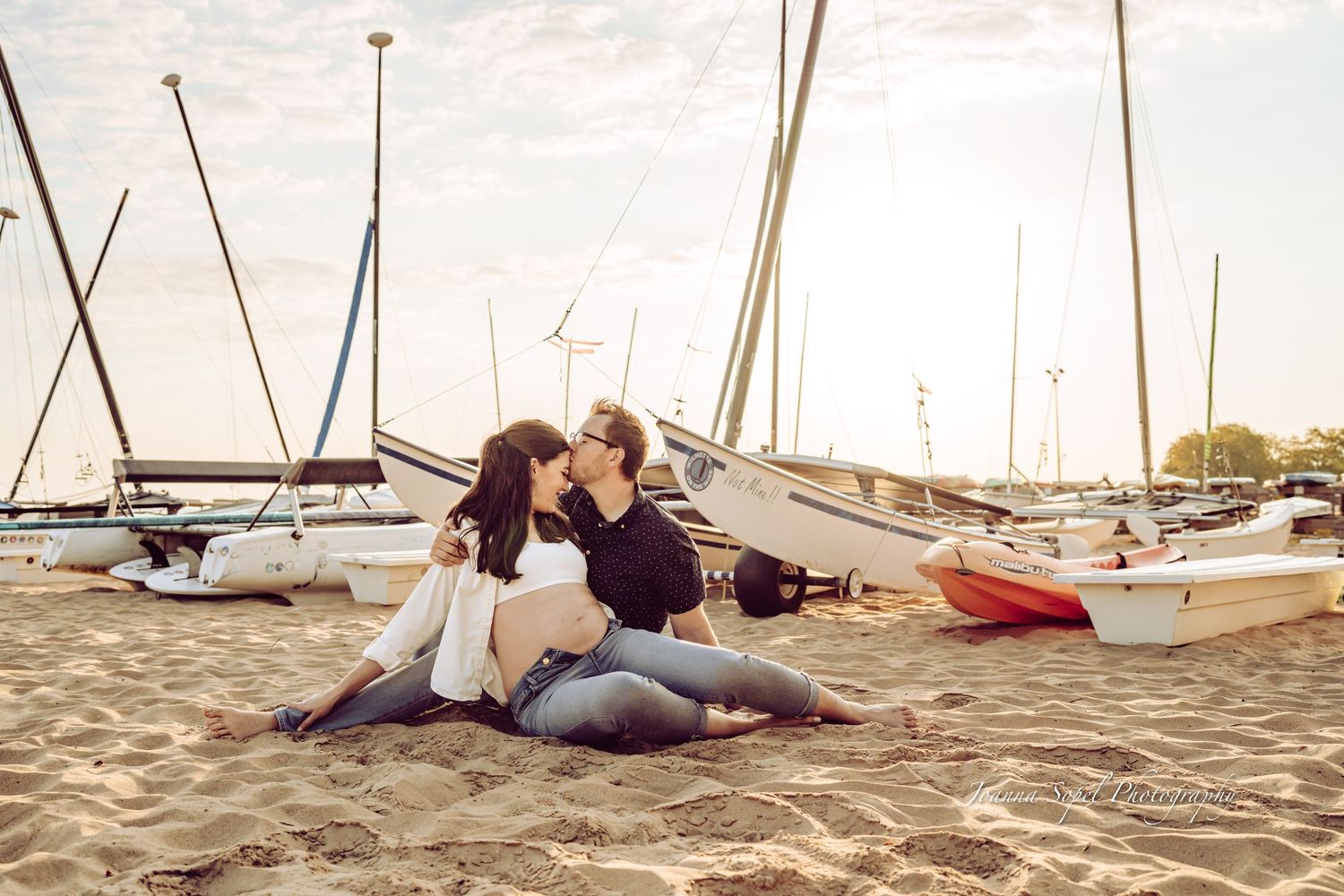 Couple sharing a kiss while sitting on sandy beach surrounded by sailboats at sunset.
