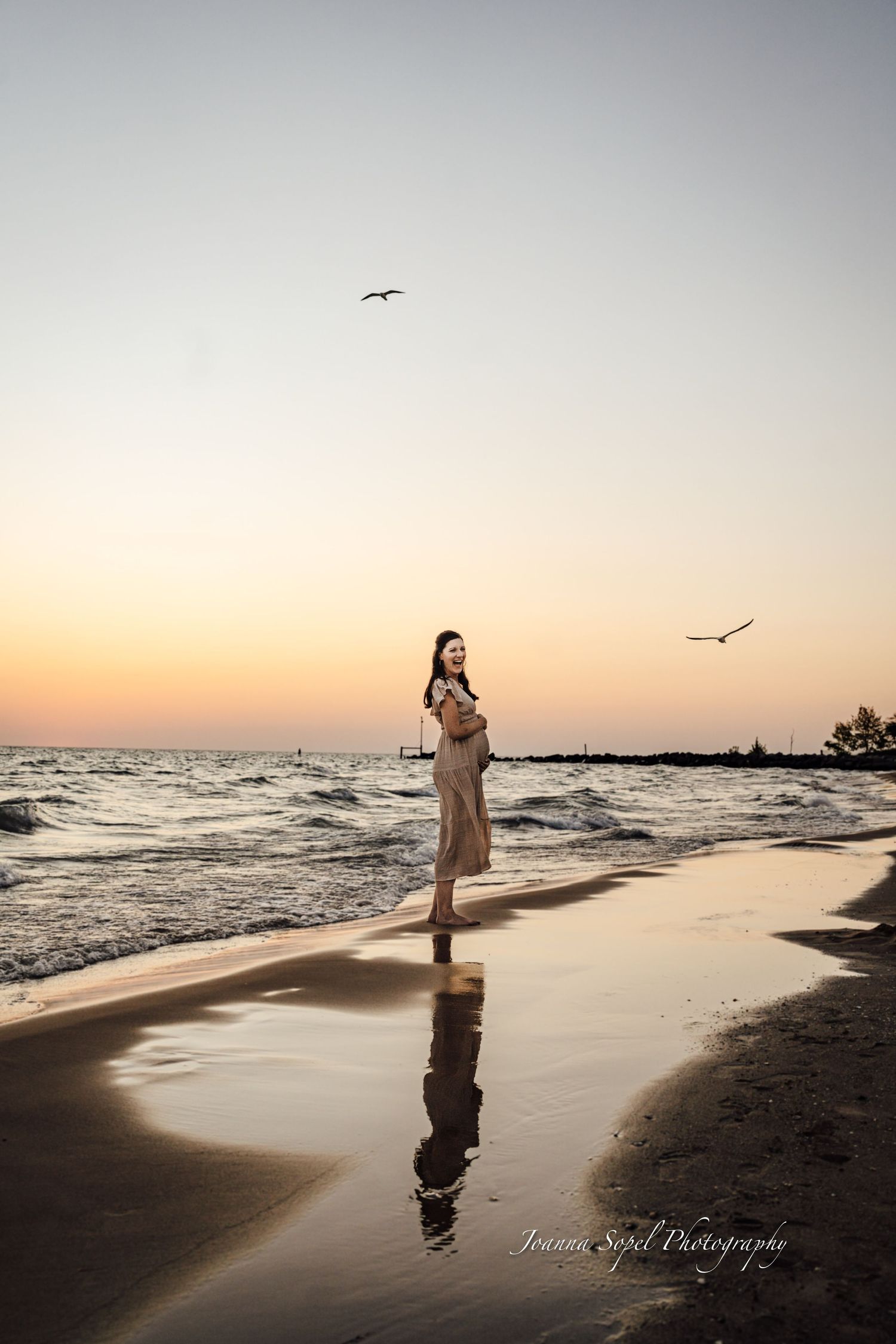 Woman in beige dress standing on sandy beach at sunset, reflected in water, with seagulls flying overhead.