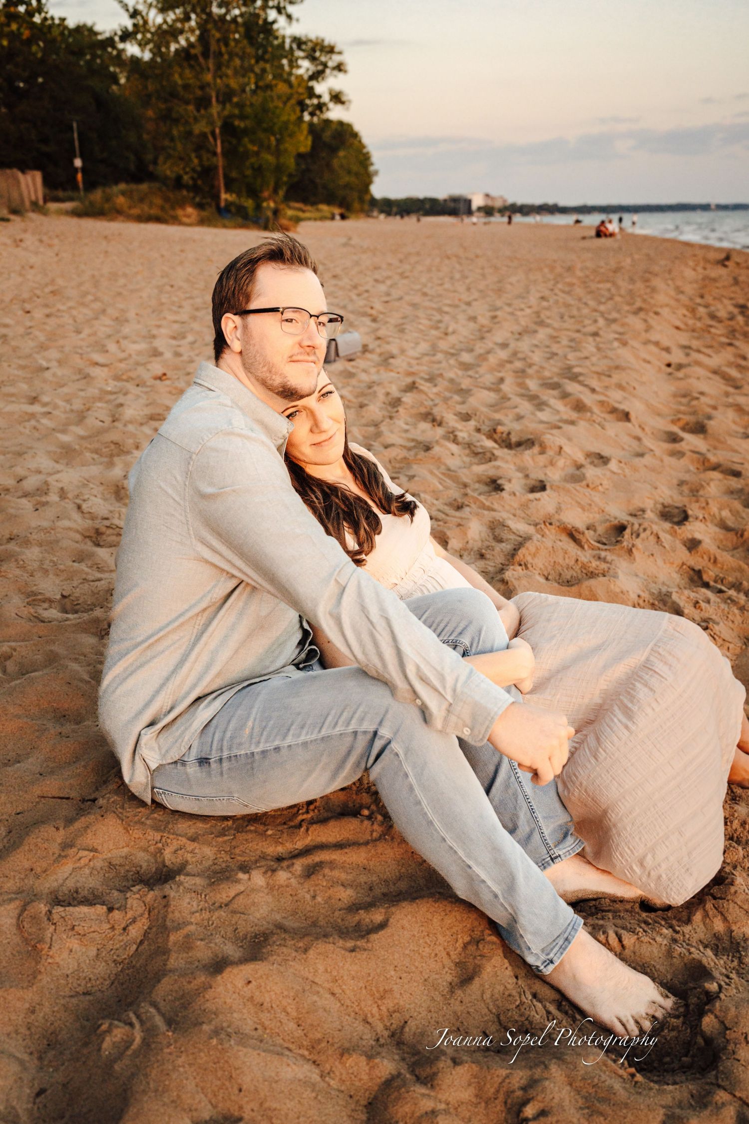 Couple sitting together on a sandy beach at sunset, woman leaning against man as he holds her close.