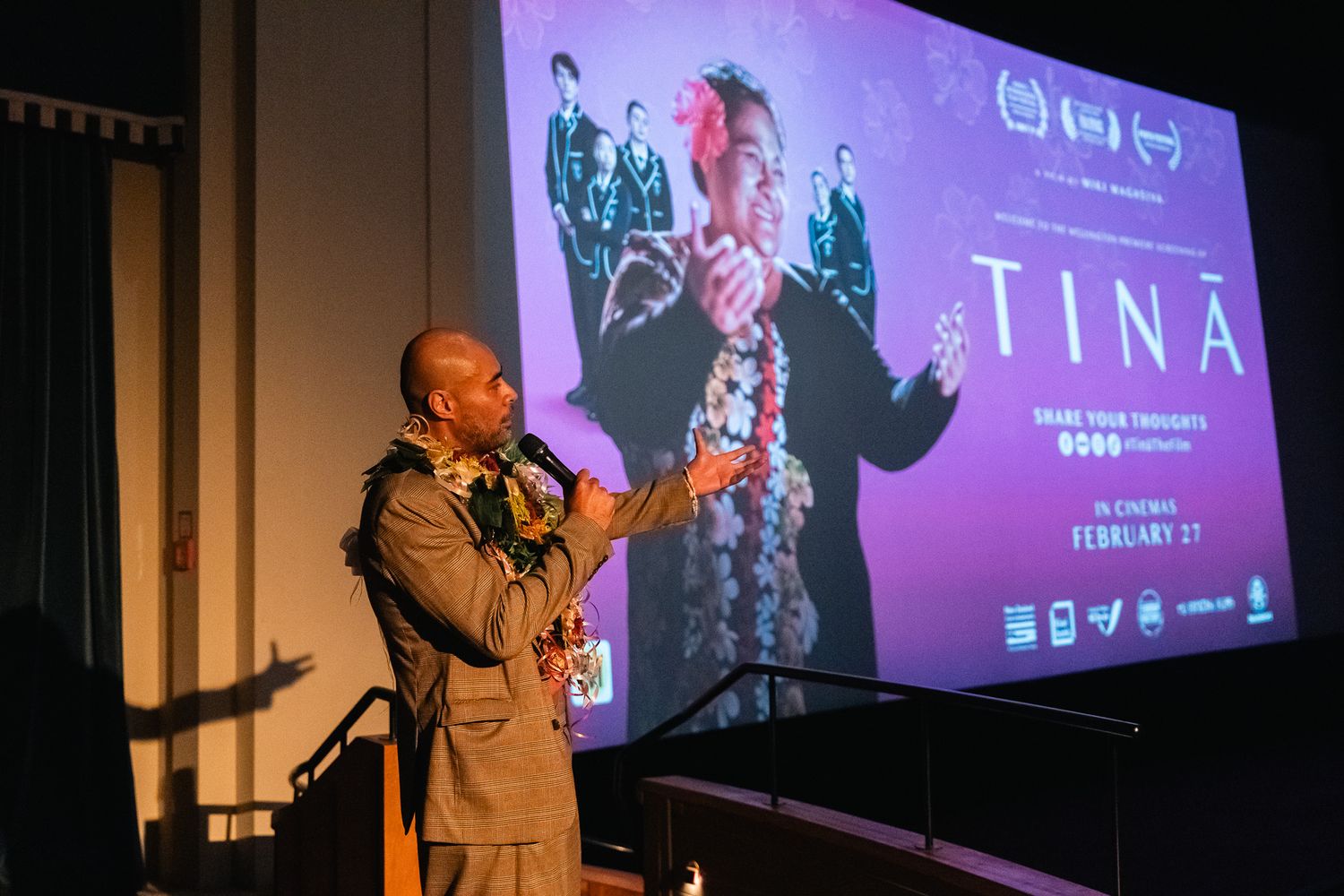 The director of Tinā gesturing to the crowd in the Embassy Theatre. The promotional slide for the film is on screen behind him.