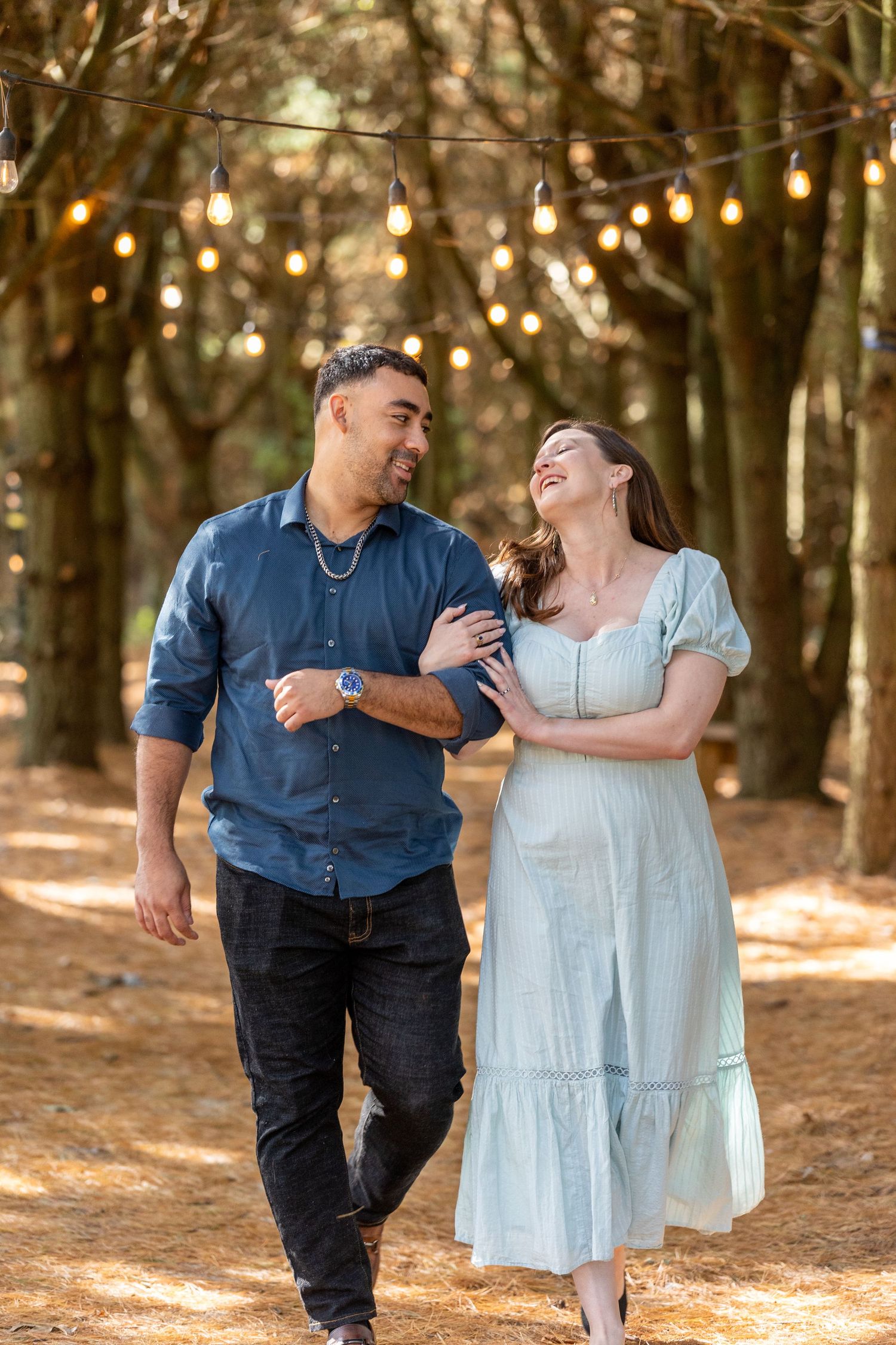 Happy couple walks arm-in-arm under string lights in a sunlit wooded outdoor setting.