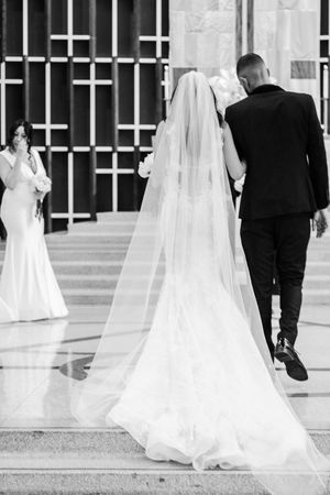 Black and white wedding photo showing bride in long veil and groom walking with bridesmaid in background.