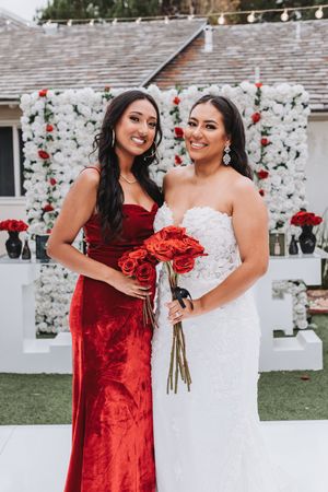Two wedding attendees share a joyful moment outdoors, with one wearing a red velvet dress and one in white bridal gown.