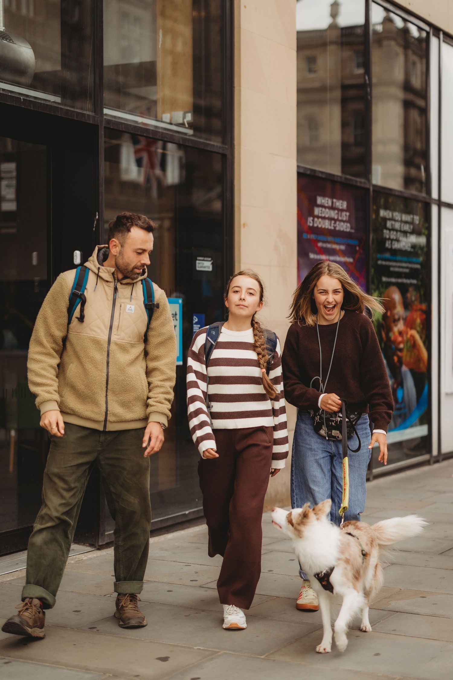 Family of three walking dog on city sidewalk, smiling and enjoying an urban stroll together.
