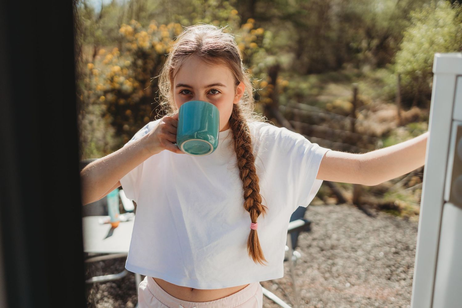 Young woman with braided hair drinking from a blue mug outdoors in a white crop top.