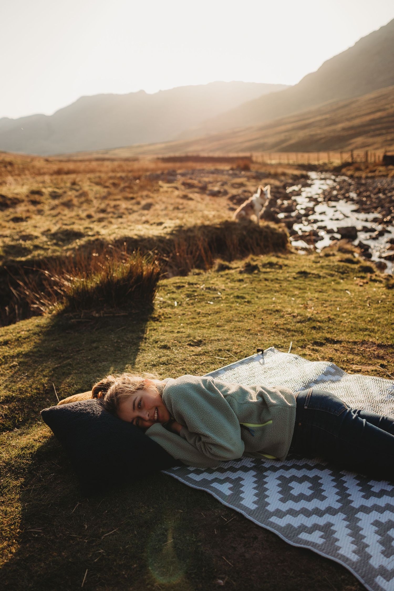 Woman lying on a blanket on green grass in a sunlit mountain valley landscape.
