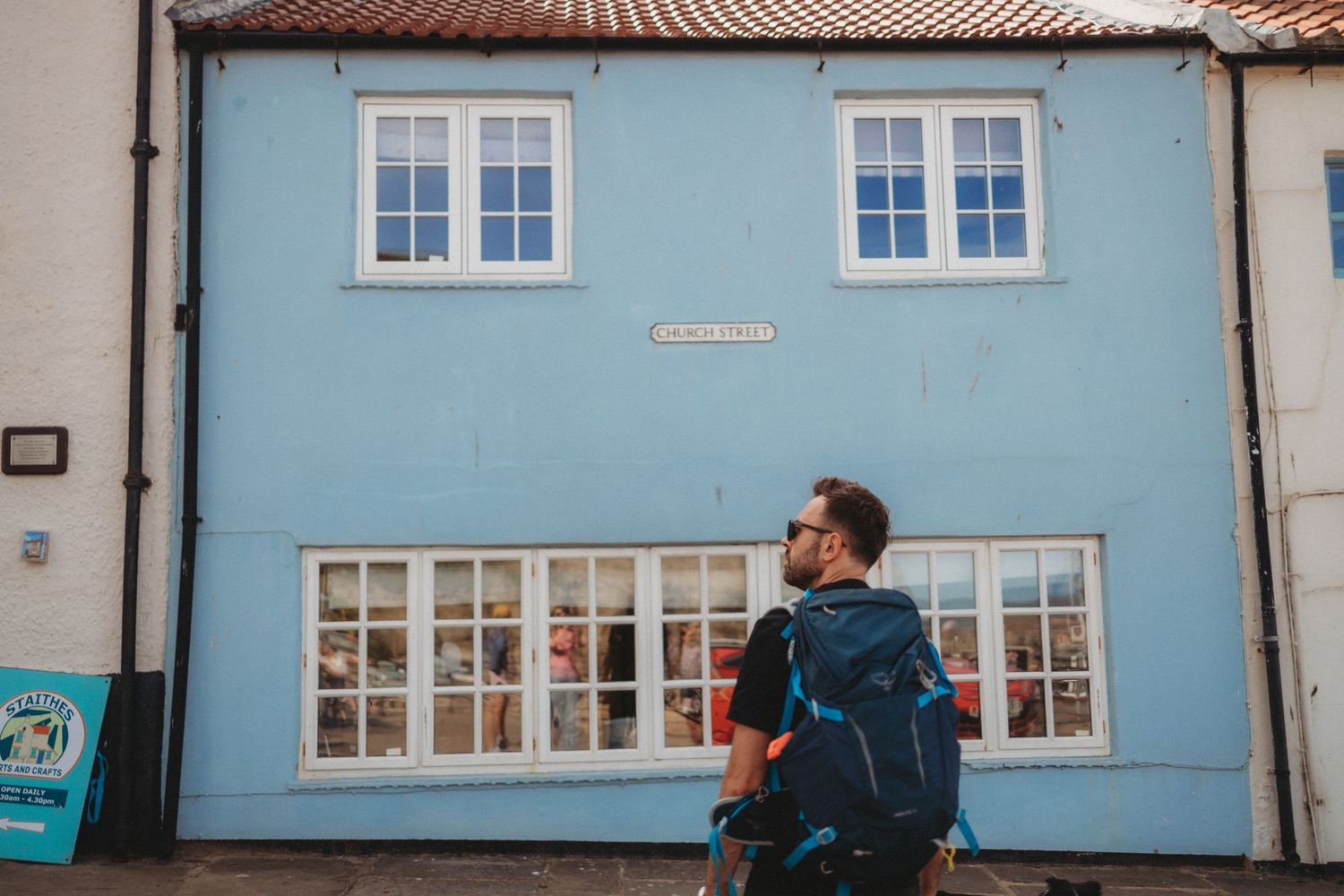 Man with backpack and sunglasses standing in front of a charming blue building with white windows.