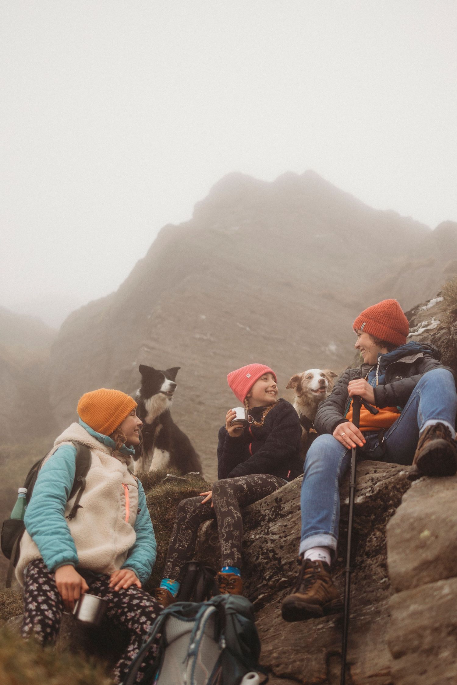 Group of hikers and their dogs taking a break on misty mountain rocks while wearing orange beanies and outdoor gear.