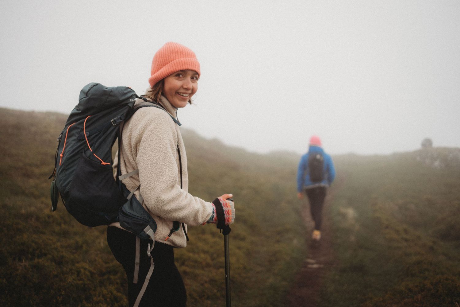 Two hikers trekking through misty mountain terrain, woman in orange beanie smiling with trekking poles and backpack.
