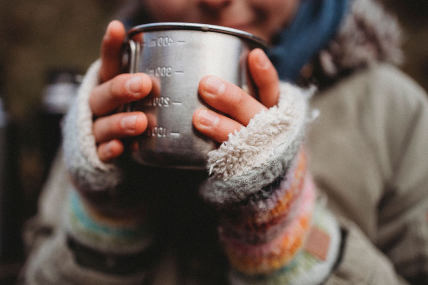 Hands in cozy sweater sleeves hold a metal camping mug with measurement markings in soft focus.