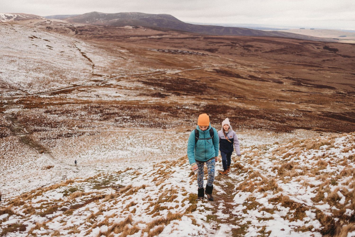 Two hikers in winter gear trek across a snowy mountain trail with brown terrain visible.