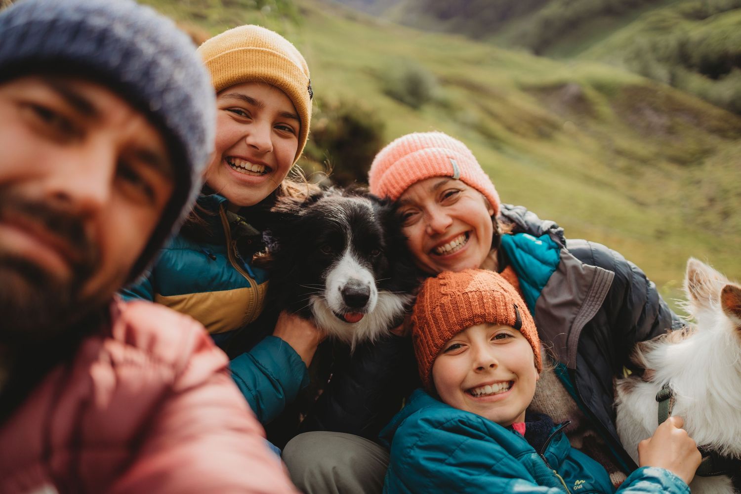 Happy family with two dogs taking a selfie outdoors in green hills, wearing colorful winter hats and jackets.
