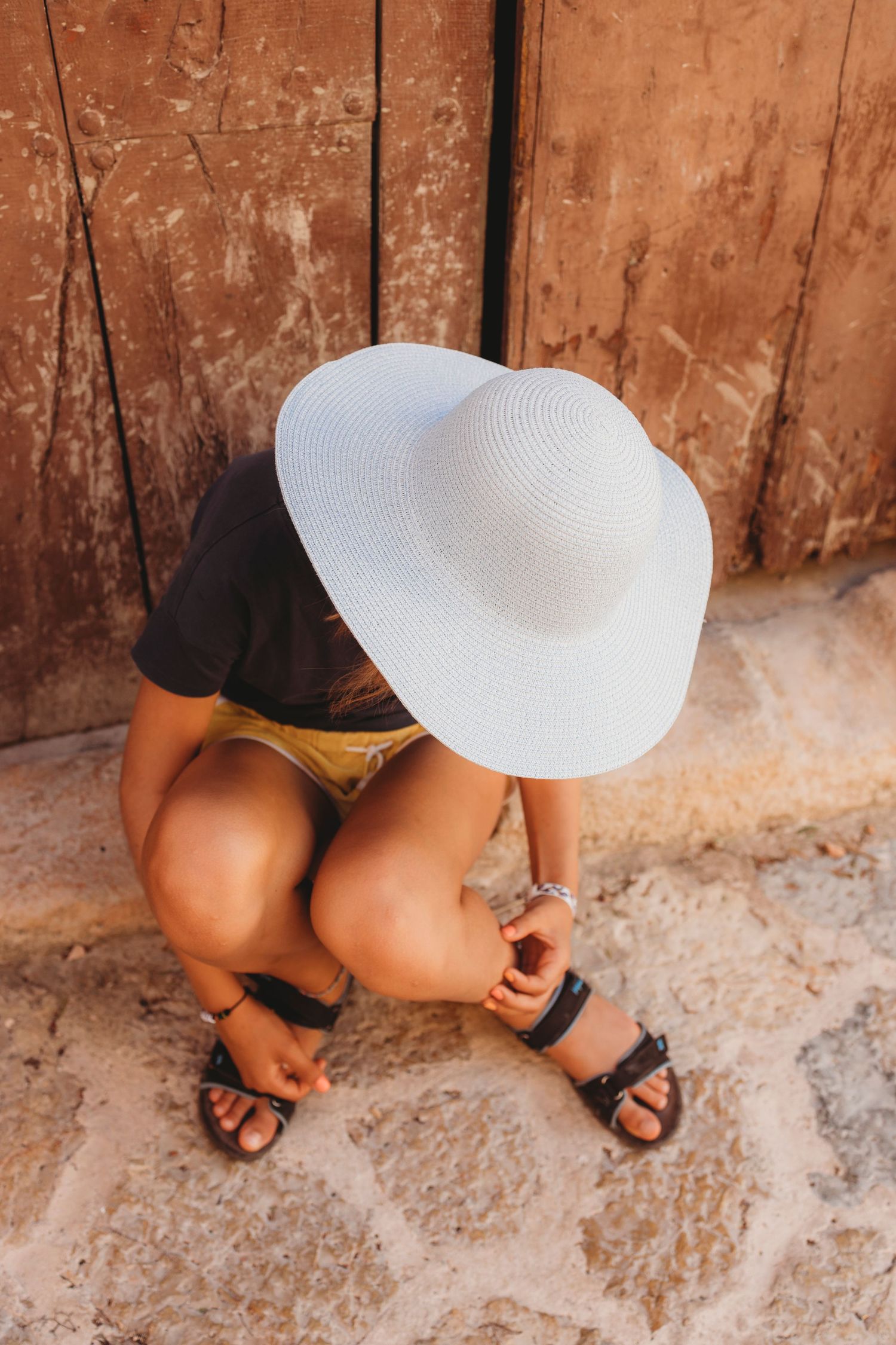 Person sitting on stone ground wearing white sun hat and sandals against rustic wooden wall.