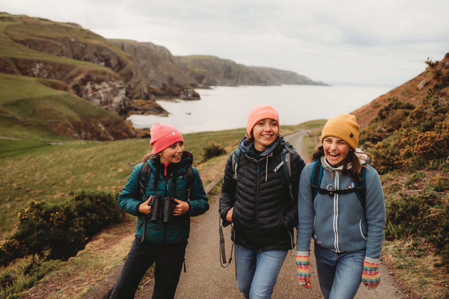 Three women hiking along a coastal path wearing colorful beanies and backpacks with scenic green cliffs and bay views.