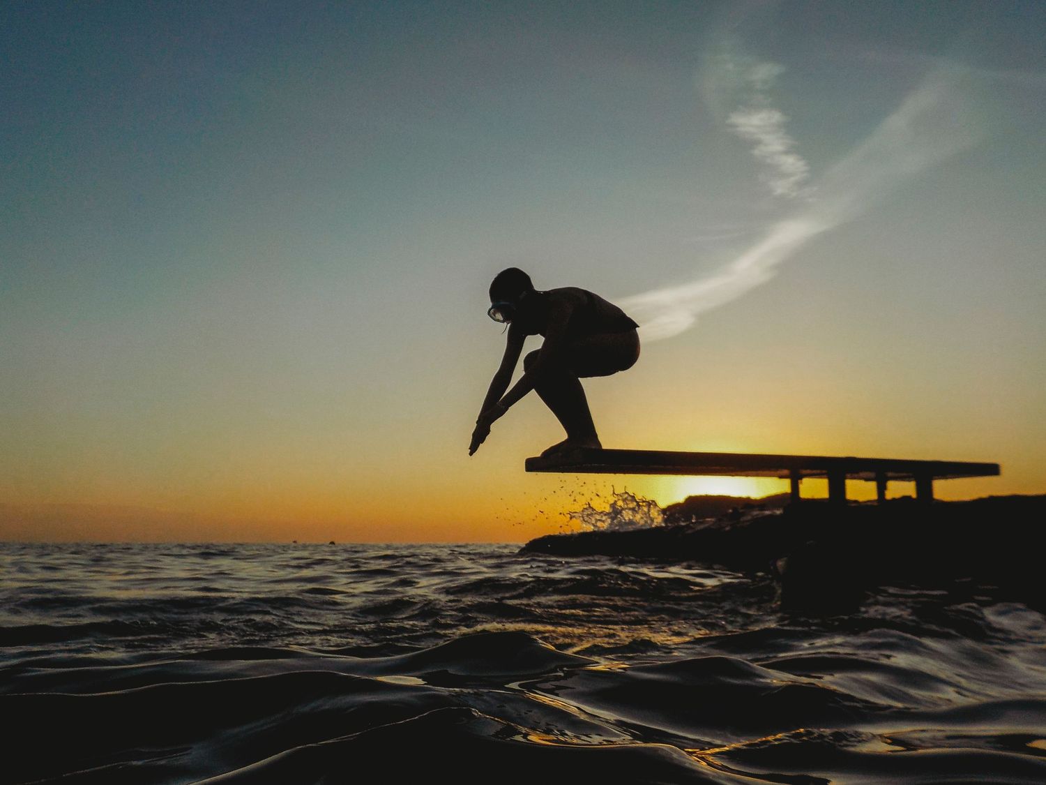 Silhouette of surfer jumping from pier at sunset over ocean waves.