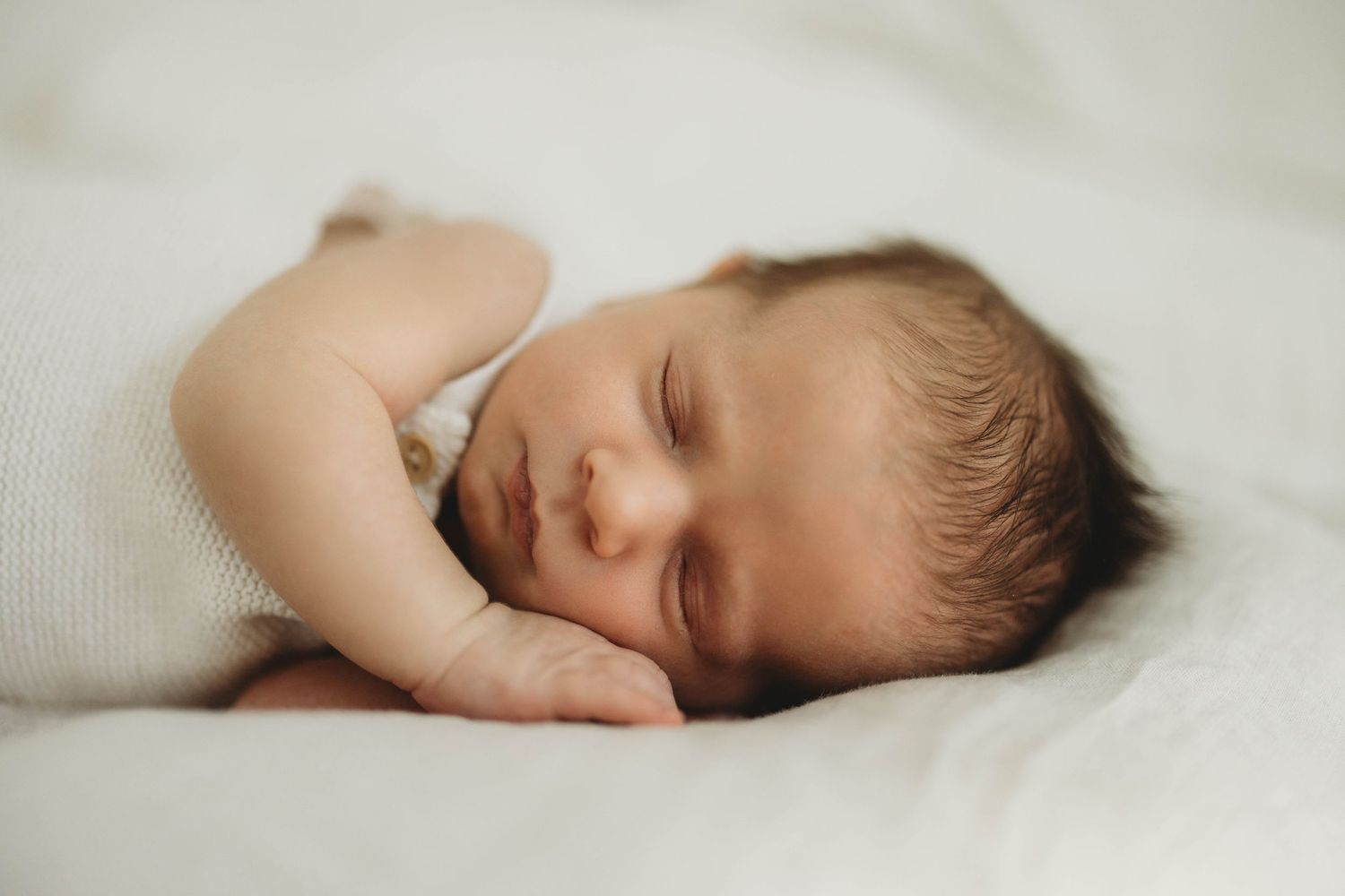 A sleeping baby lies peacefully under soft white bedding with wispy hair visible above their face.
