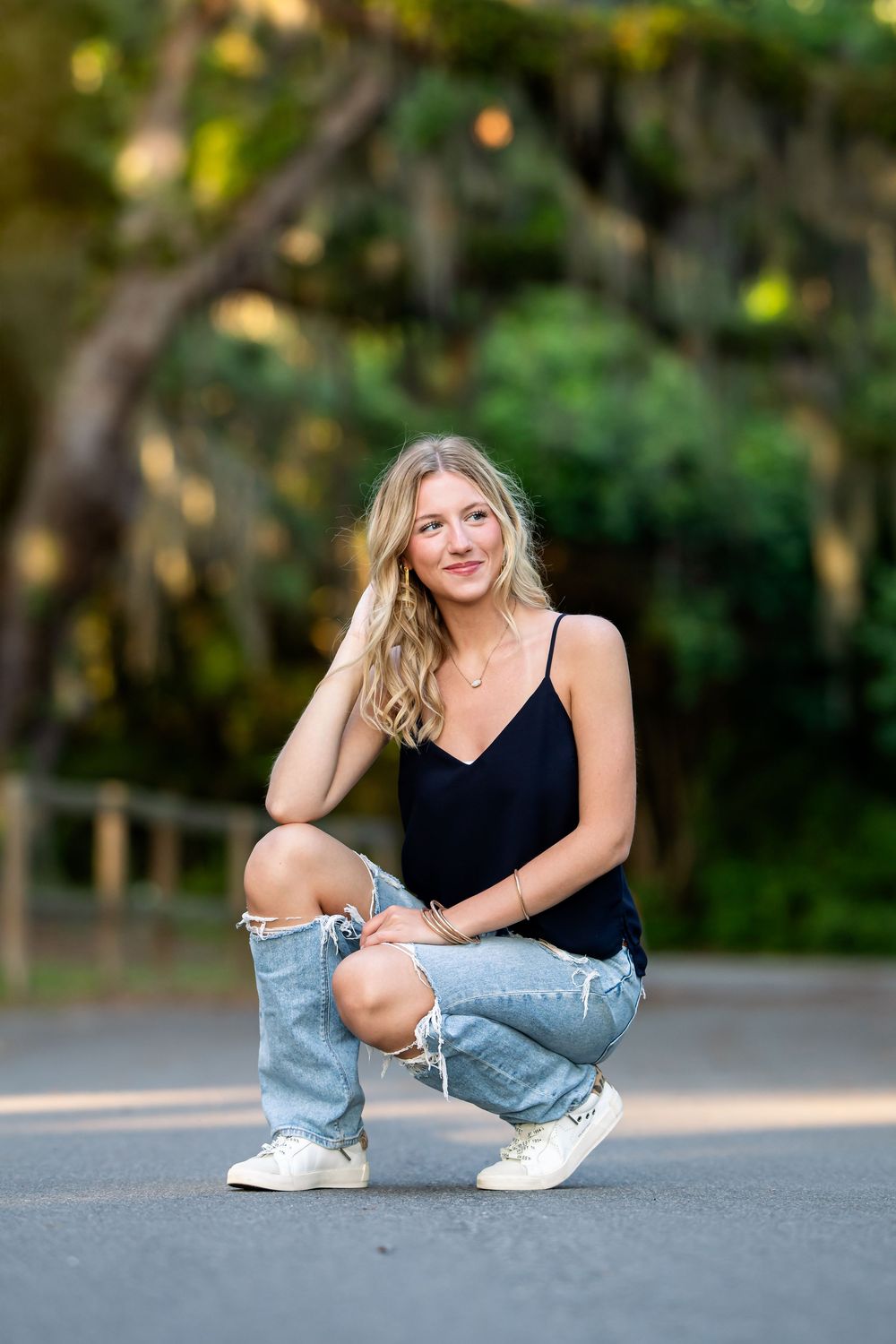A person in a black tank top and ripped jeans crouches on a paved road with trees in the background.