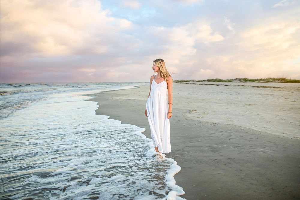 A person in a flowing white dress stands at the edge of the ocean waves during a dreamy golden hour sunset.