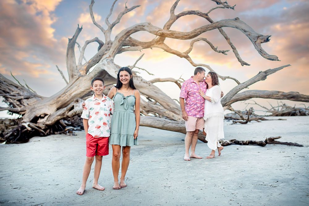 Children play near a dramatic driftwood tree on a beach during sunset with pink clouds in the background.