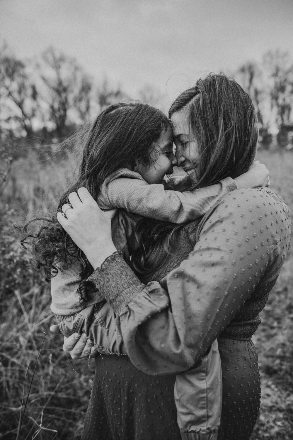 Intimate black and white portrait of a couple embracing outdoors in a field with trees in the background.