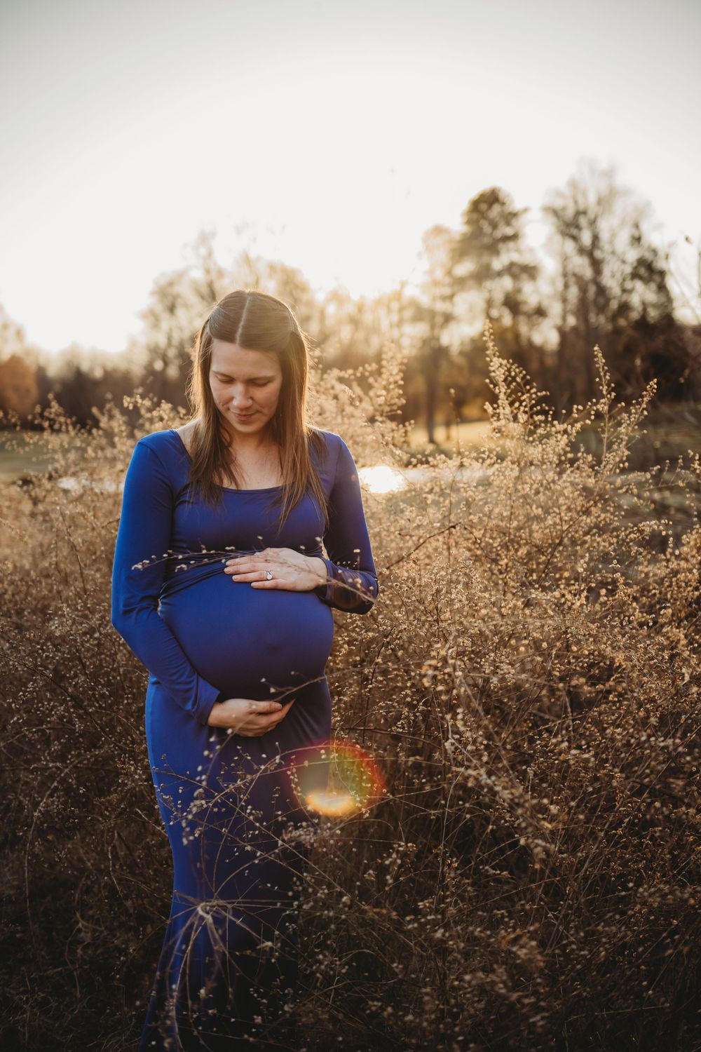A maternity photoshoot in a golden field at sunset captures a serene moment with a woman in a blue dress.