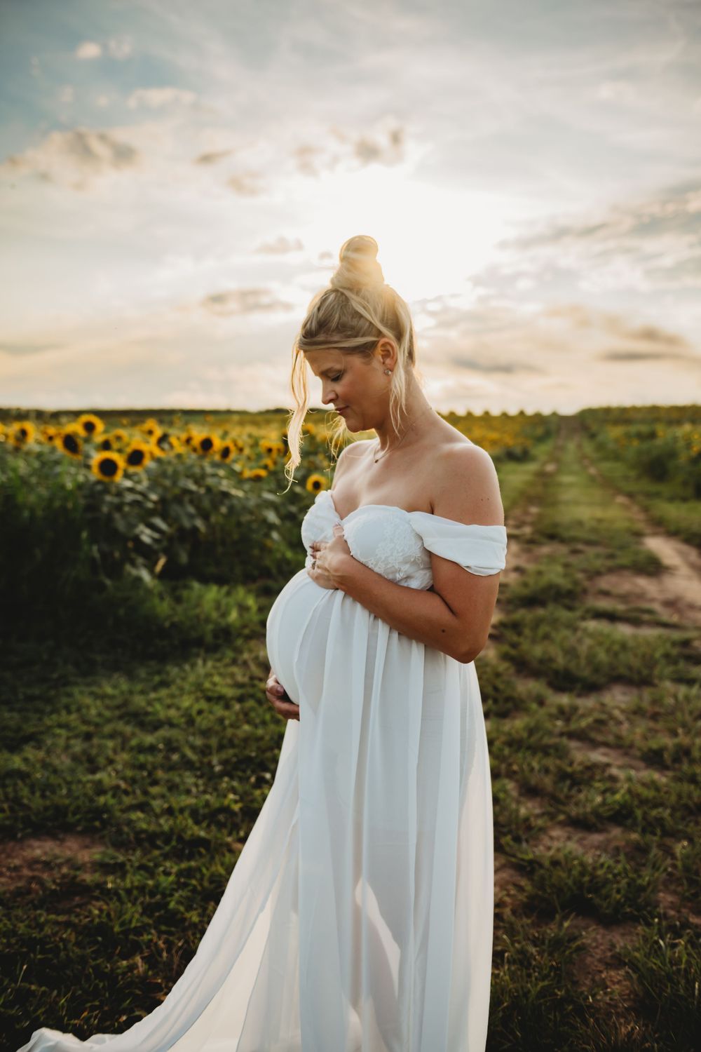 Ethereal maternity photoshoot in a sunlit sunflower field at golden hour with flowing white dress and glowing sunset.