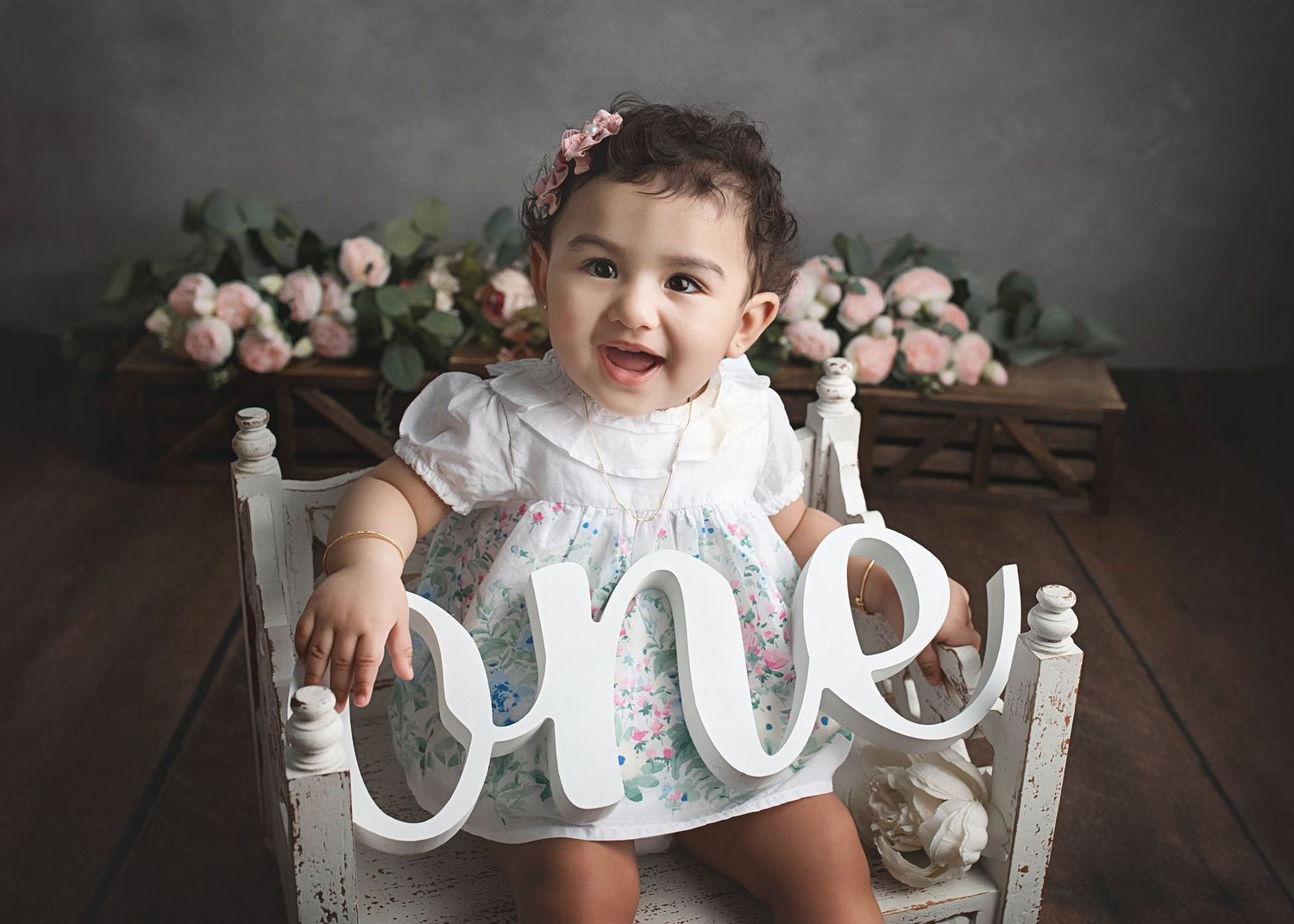 A child sits in a white vintage chair with floral decor, holding a decorative 'ONE' sign during a milestone photoshoot.
