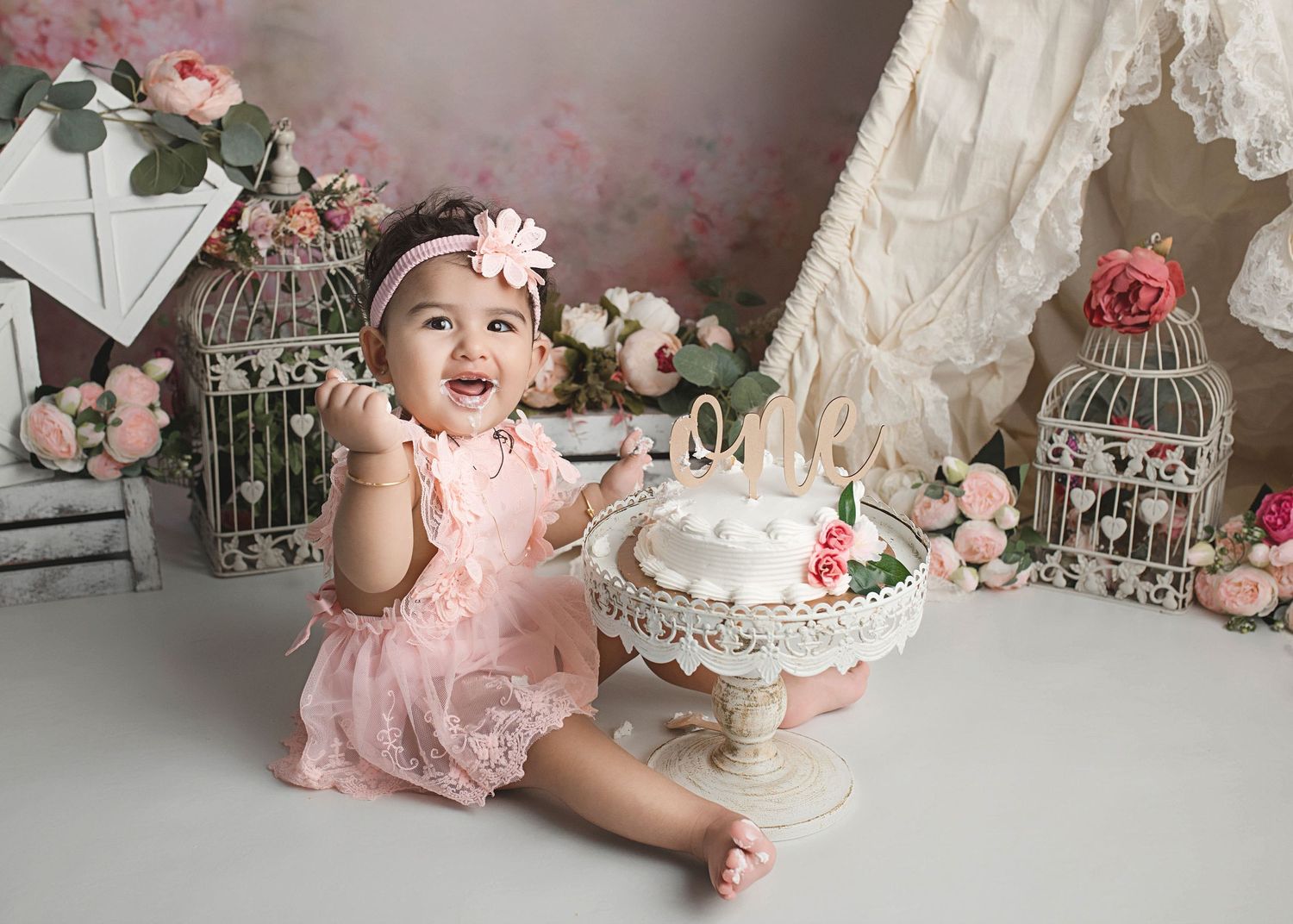 Vintage-themed cake smash photo setup with decorative birdcages, pink roses, lace curtains and ornate white cake stand.