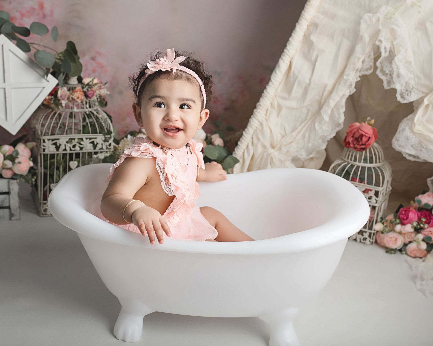 A smiling baby sits in a white clawfoot bathtub surrounded by pink roses and decorative birdcages during a photo shoot.