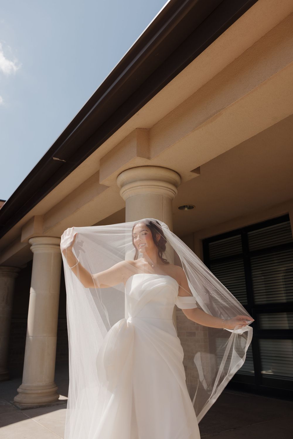 Elegant white wedding dress with flowing veil captured under a portico with classical columns in natural daylight.