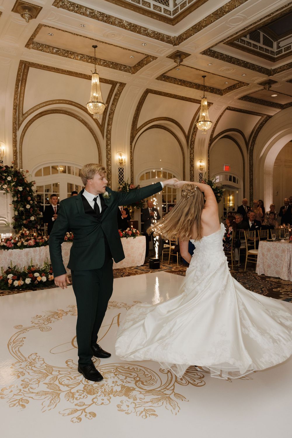 Couple dancing in an elegant ballroom with ornate ceiling and decorative floor during their first dance at a wedding reception.