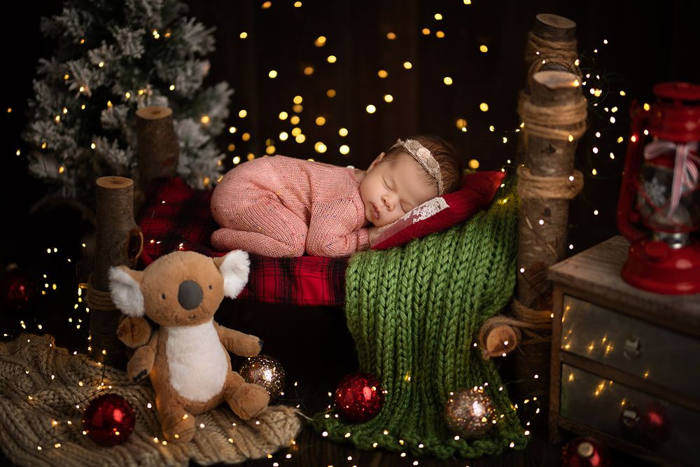 A sleeping baby in red pajamas rests on a green blanket surrounded by Christmas decorations and twinkling lights.