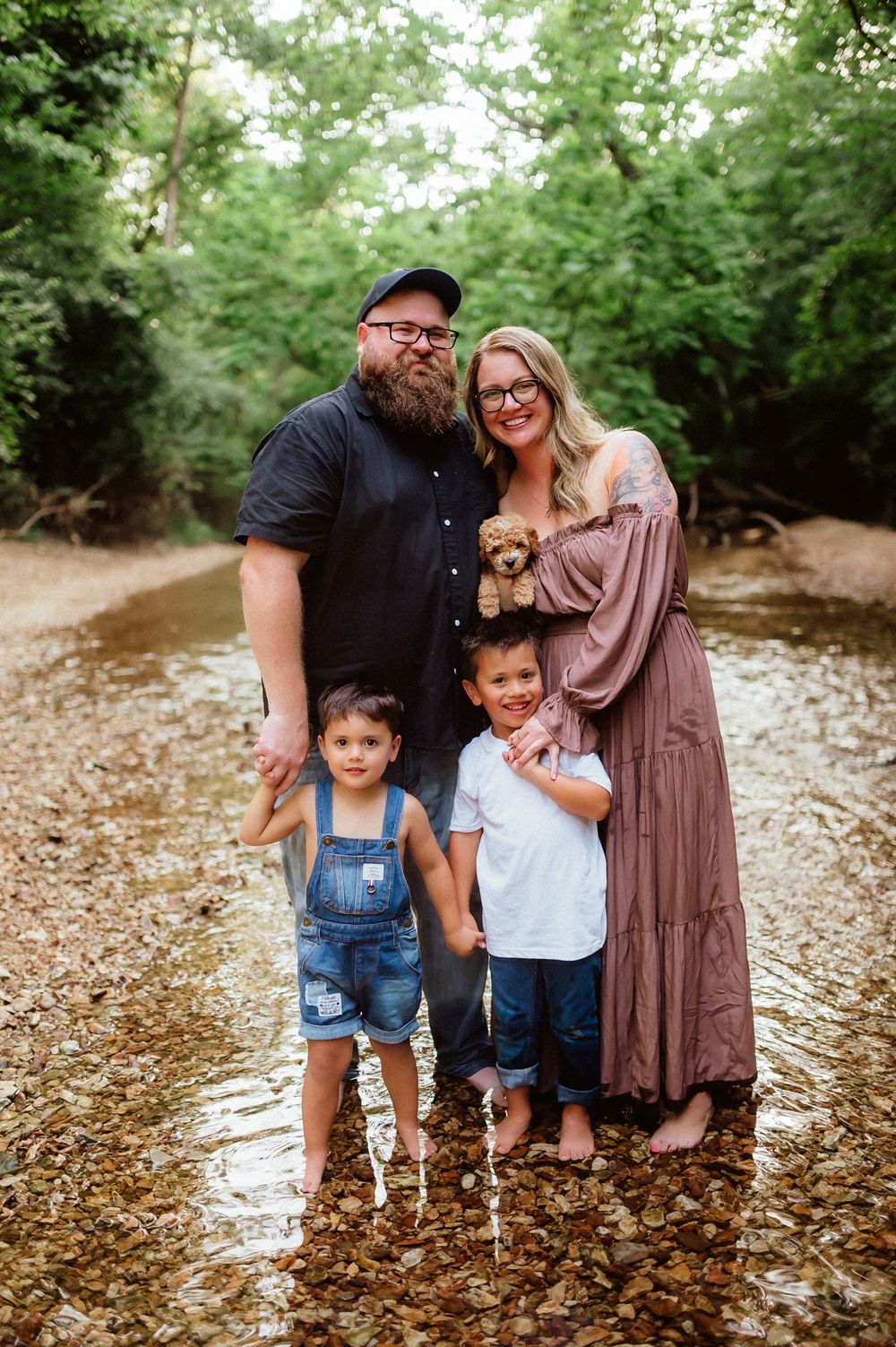A family stands together on fallen autumn leaves in a wooded outdoor setting for a casual photo session.