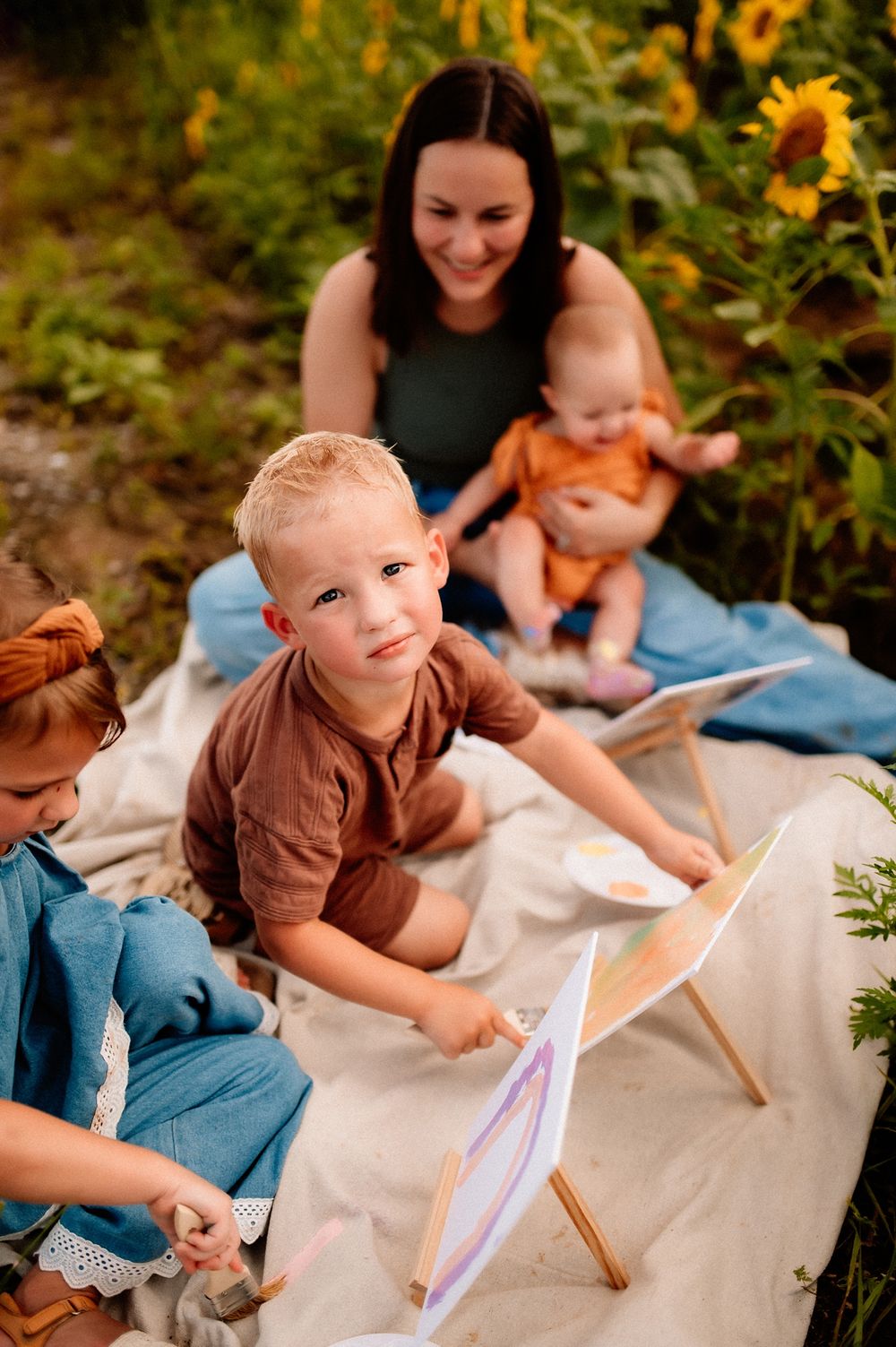 A family sits on a blanket near blooming sunflowers while children draw and play together on a sunny day.