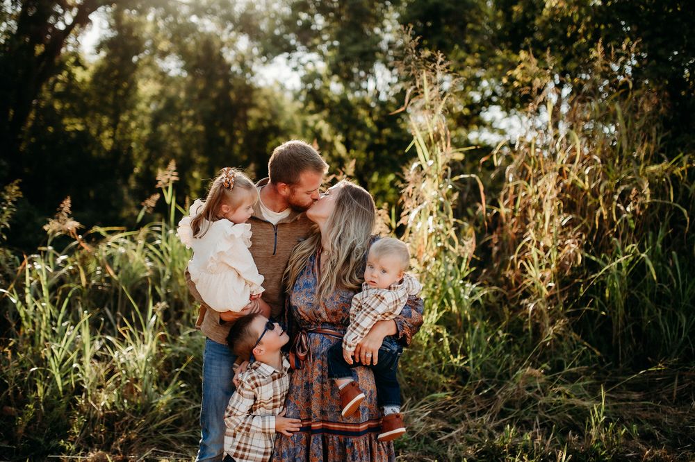 A romantic couple embraces in a sun-drenched field during golden hour with tall grasses and trees in the background.