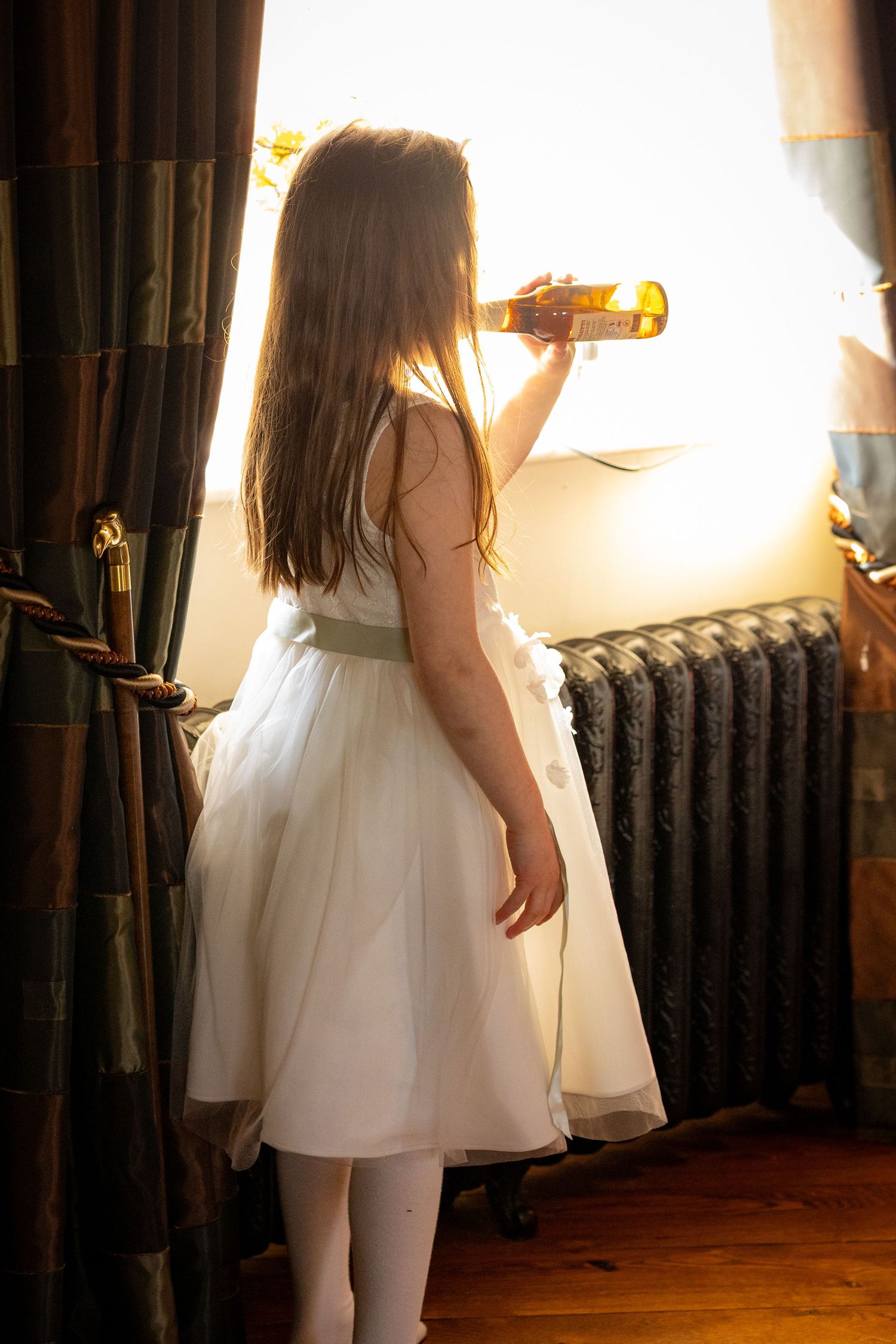 Flower girl standing by a window pretending to drink from a beer bottle inside the windmill at Windmill Farm in Burscough.