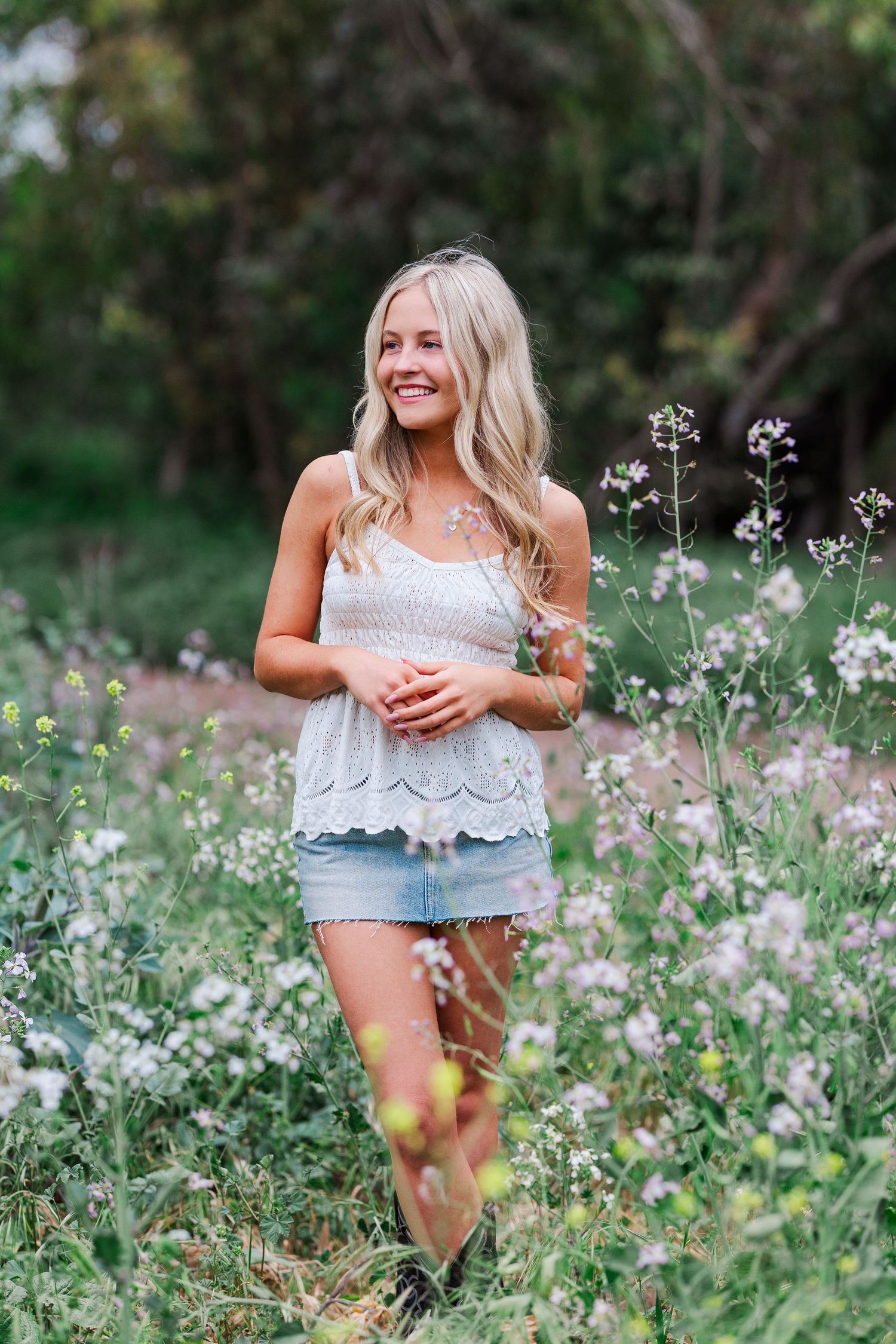 A person in a white top and denim shorts poses playfully among blooming wildflowers in a lush garden setting.