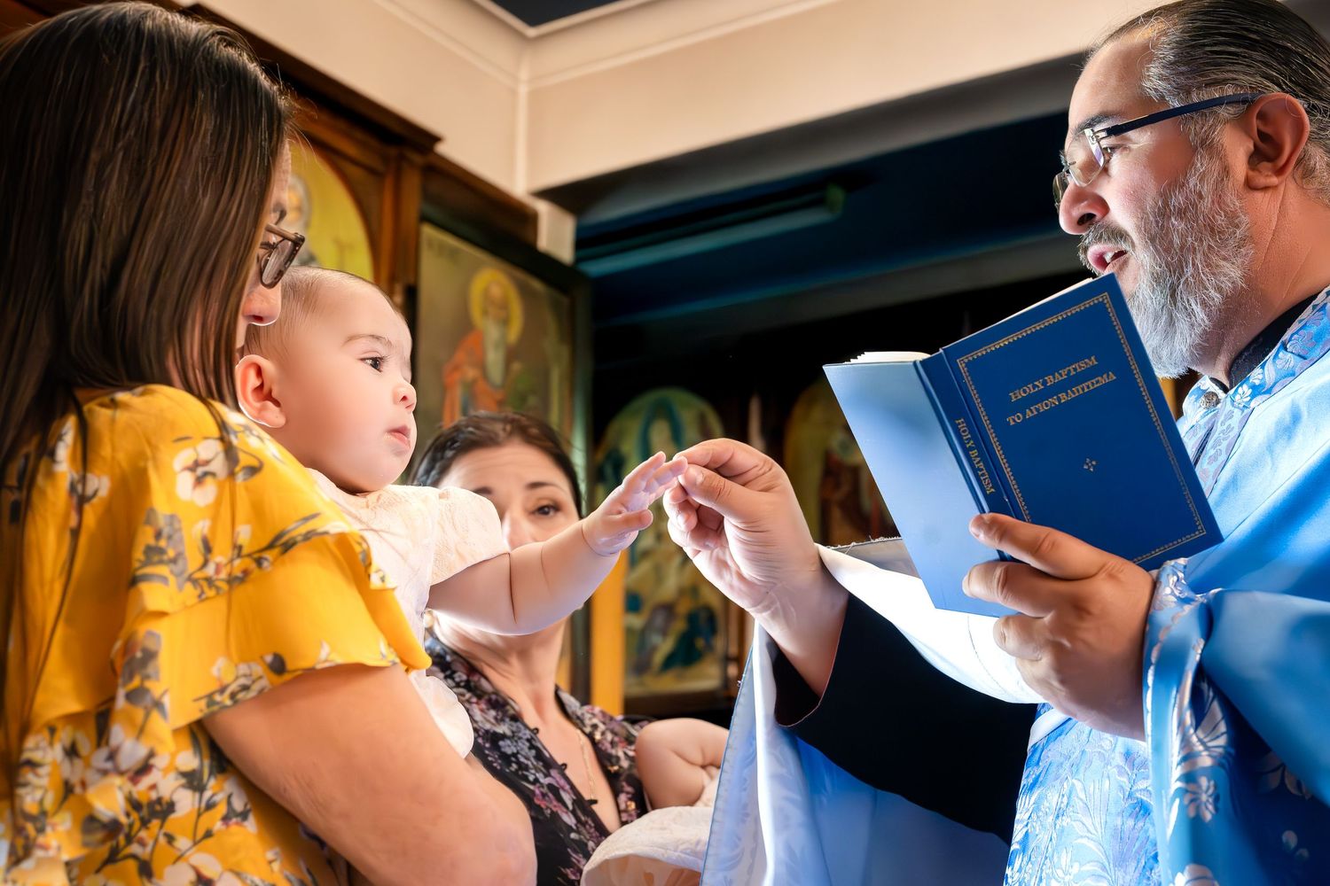 Orthodox priest in blue vestments reads from holy book during infant baptism ceremony with mother holding baby.