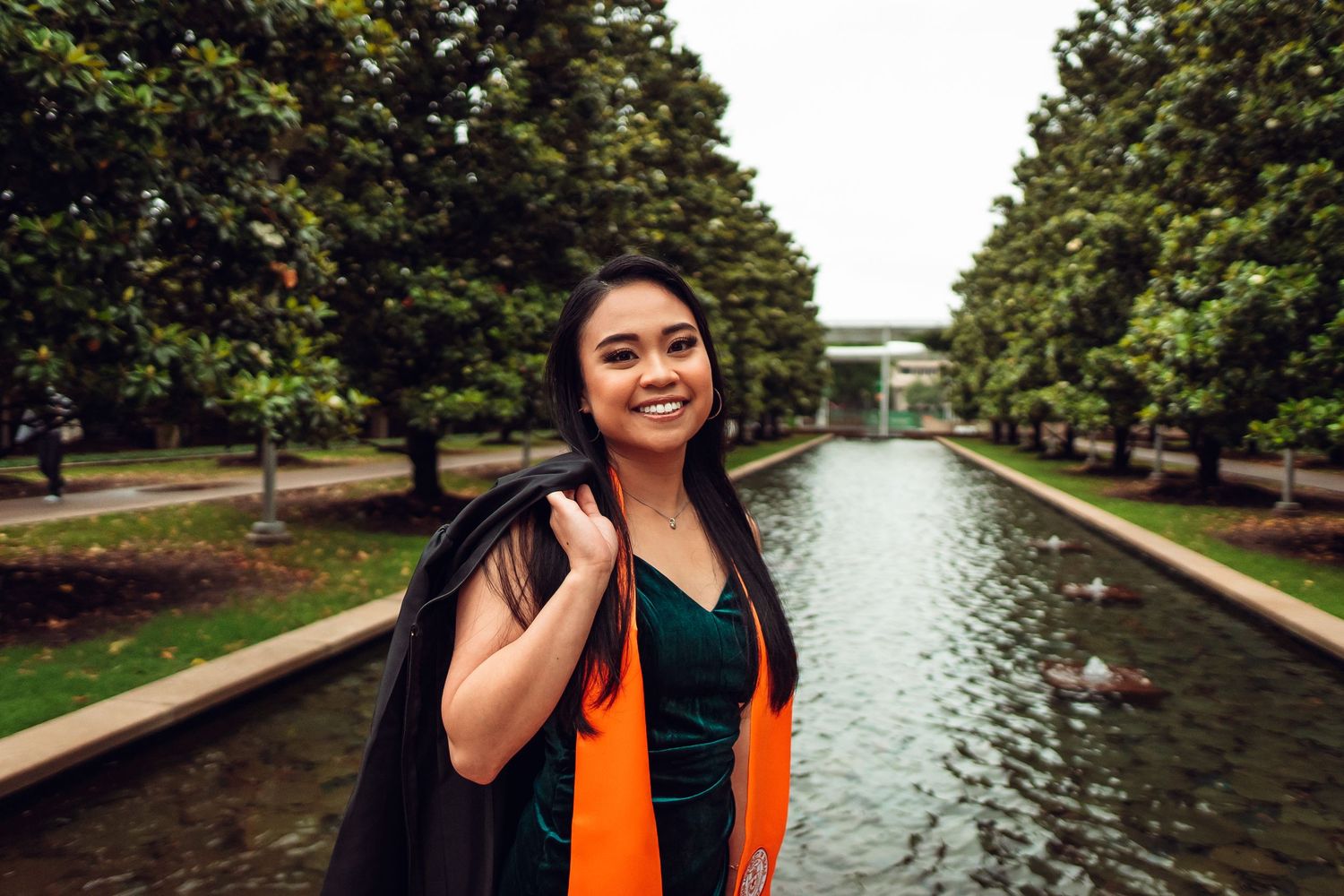 Graduate in orange stole walking along a scenic campus waterway surrounded by trees.