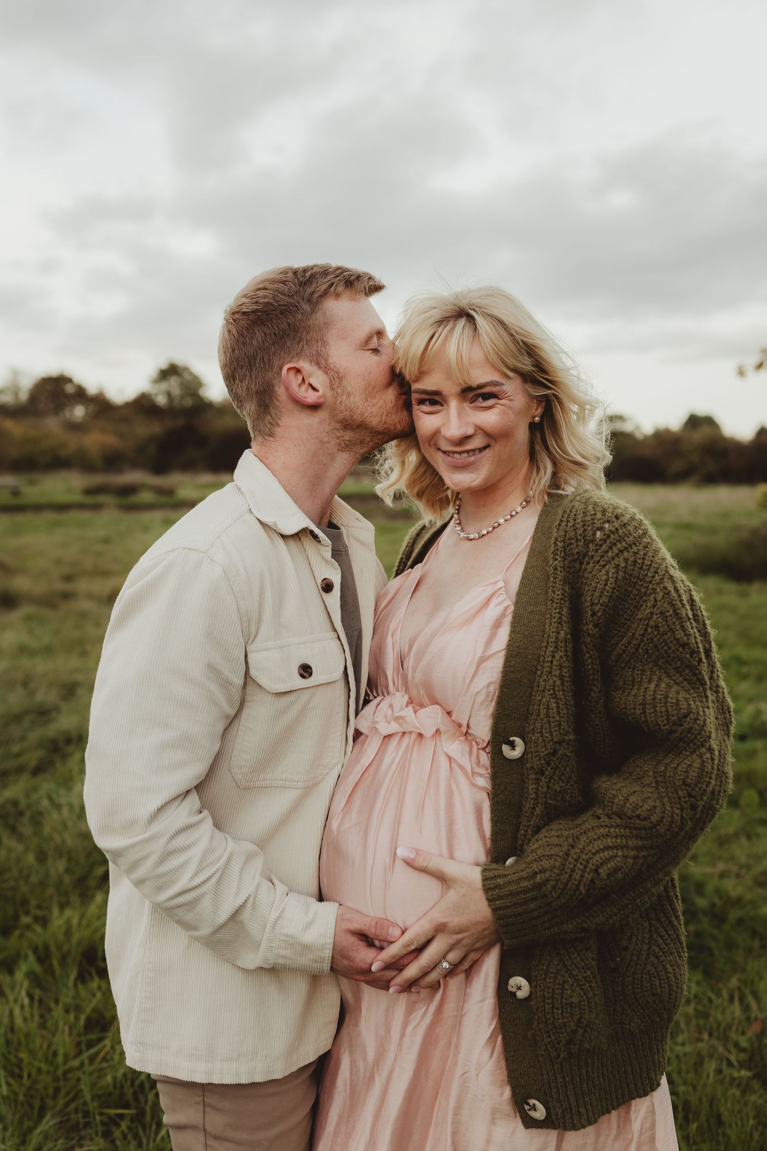 A romantic couple shares a tender kiss while holding hands, wearing neutral toned sweaters in a soft outdoor setting.