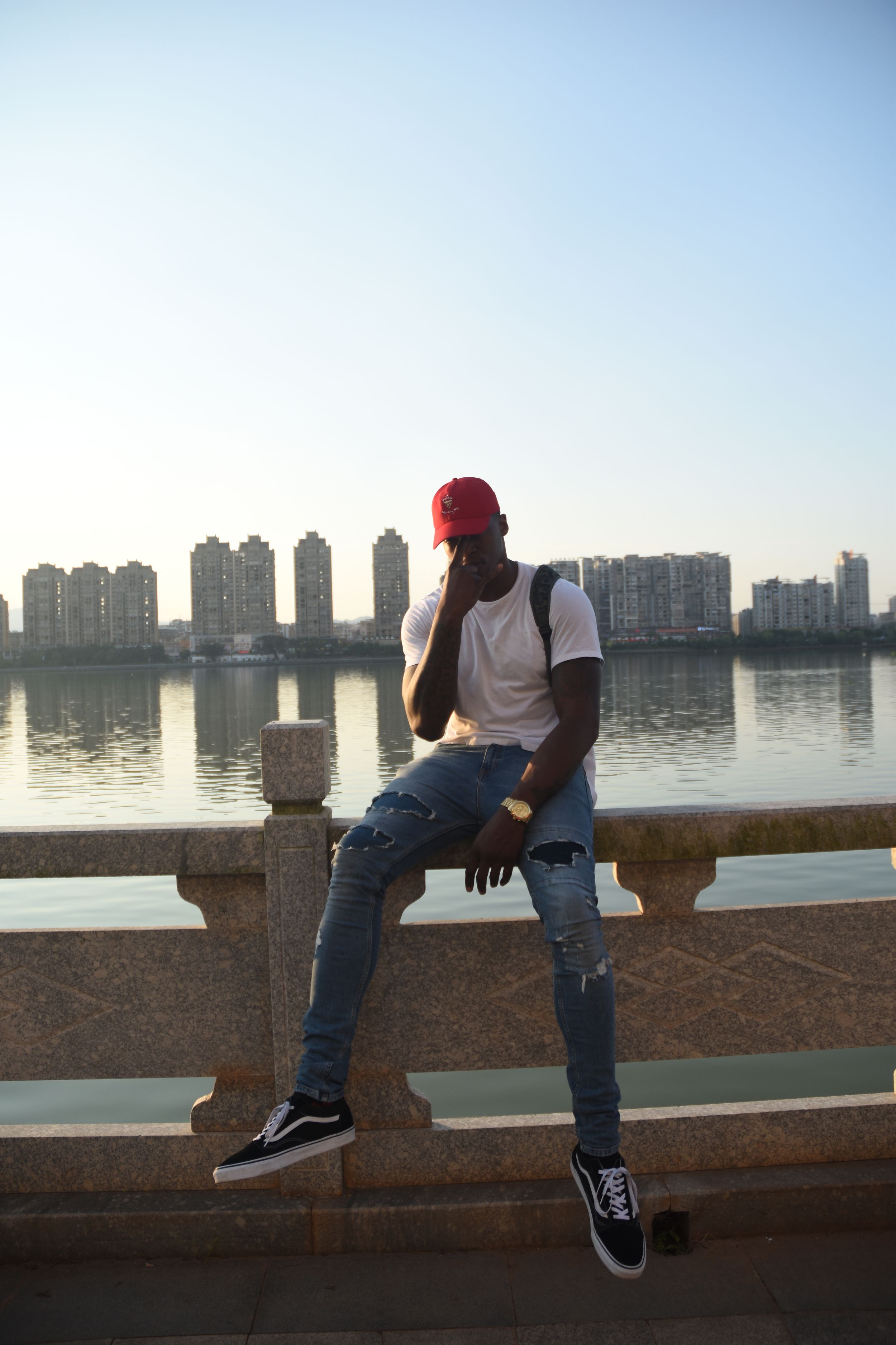 Person in red cap and casual wear sits on waterfront railing with city skyline and reflective water in background.