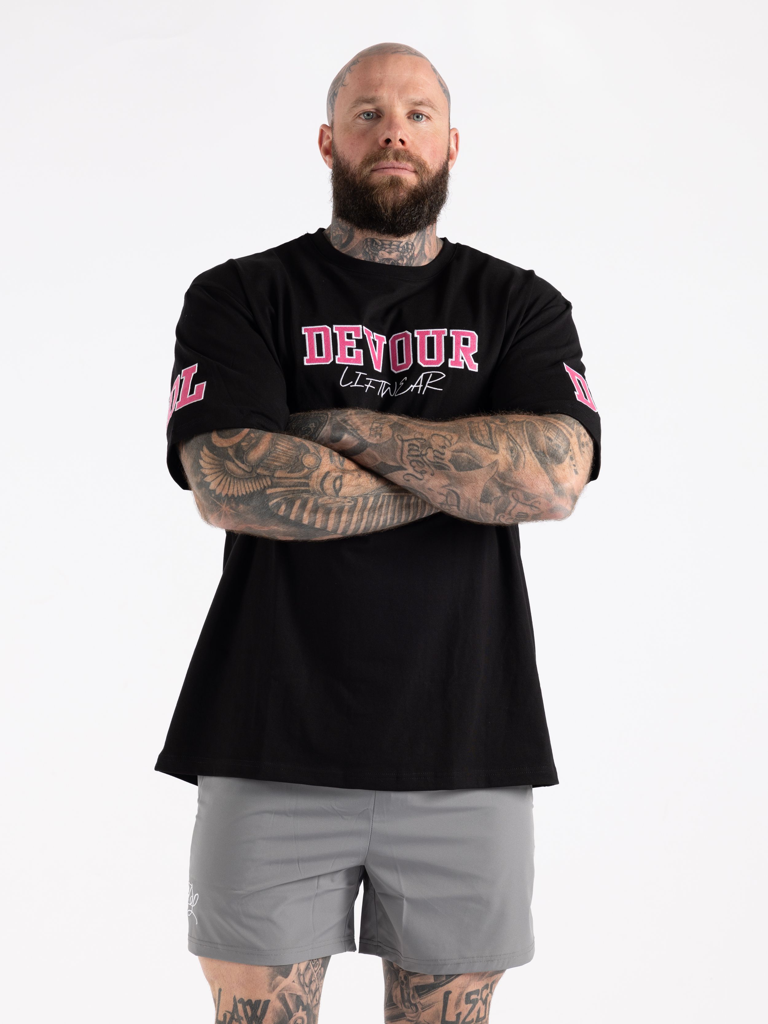 Athletic model wearing black graphic t-shirt and grey shorts shows off arm tattoos against white background.