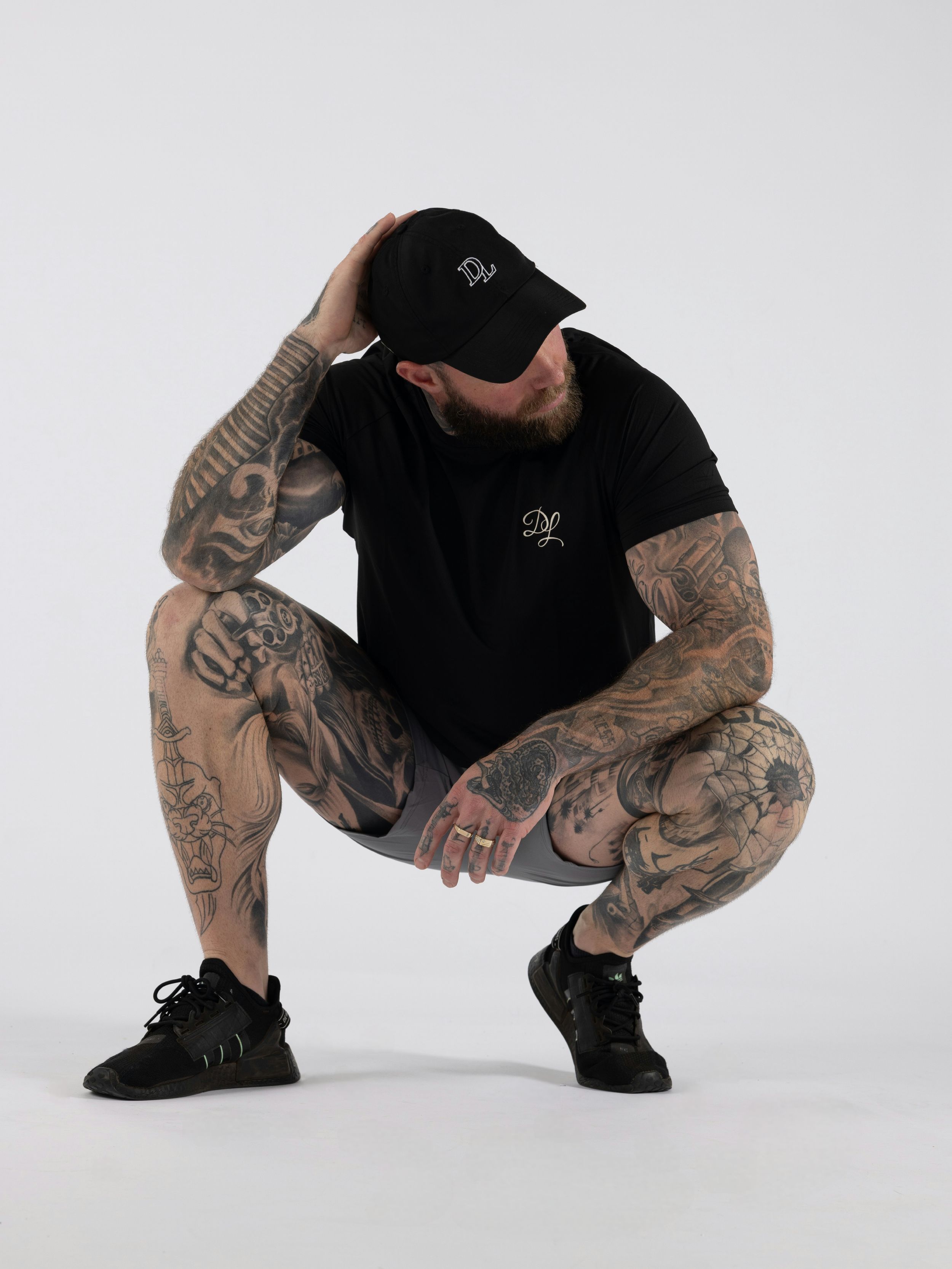 Tattooed person in black t-shirt and shorts crouching down against white background wearing black cap and sneakers.